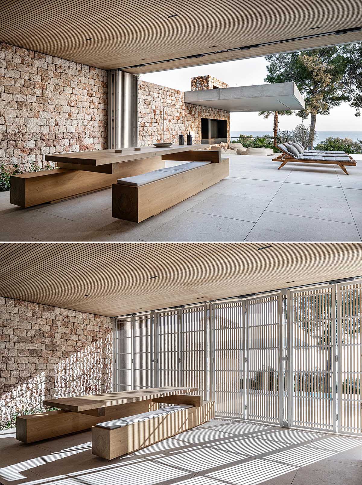 This modern covered dining area is a furnished with a custom large wood table with benches. The stone wall from the kitchen also seamlessly flows through to the outdoor areas.