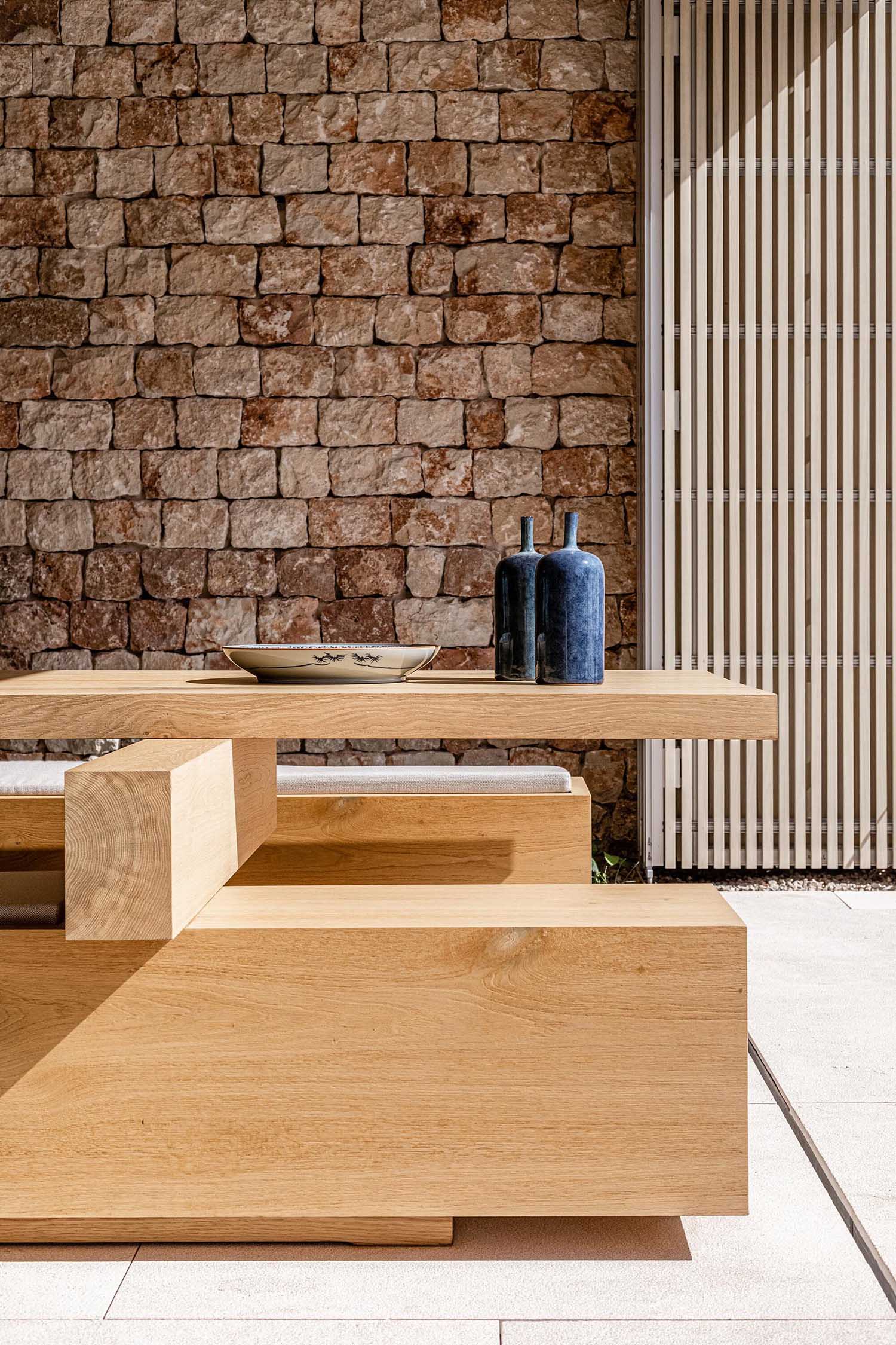 This modern covered dining area is a furnished with a custom large wood table with benches. The stone wall from the kitchen also seamlessly flows through to the outdoor areas.