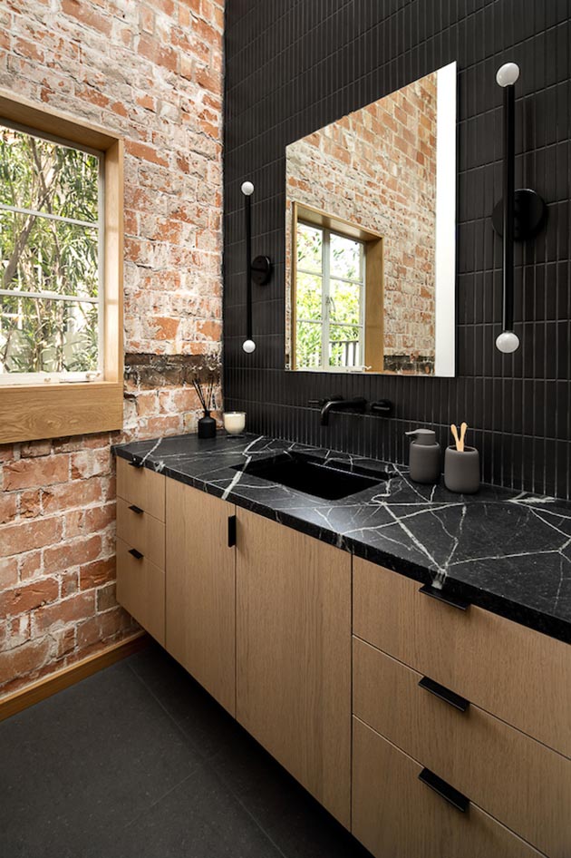 In this modern bathroom there's black wall tiles and sconces, the original brick walls, and wood vanity.