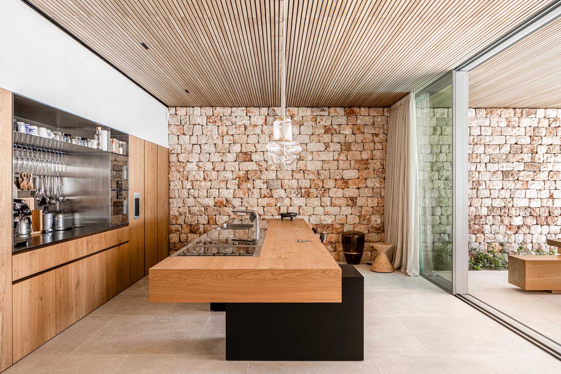 This modern kitchen includes flush wood cabinets, stainless steel lined alcove, and a large island with a thick wood countertop and black base. Sliding glass doors open the kitchen up to a covered outdoor dining area.