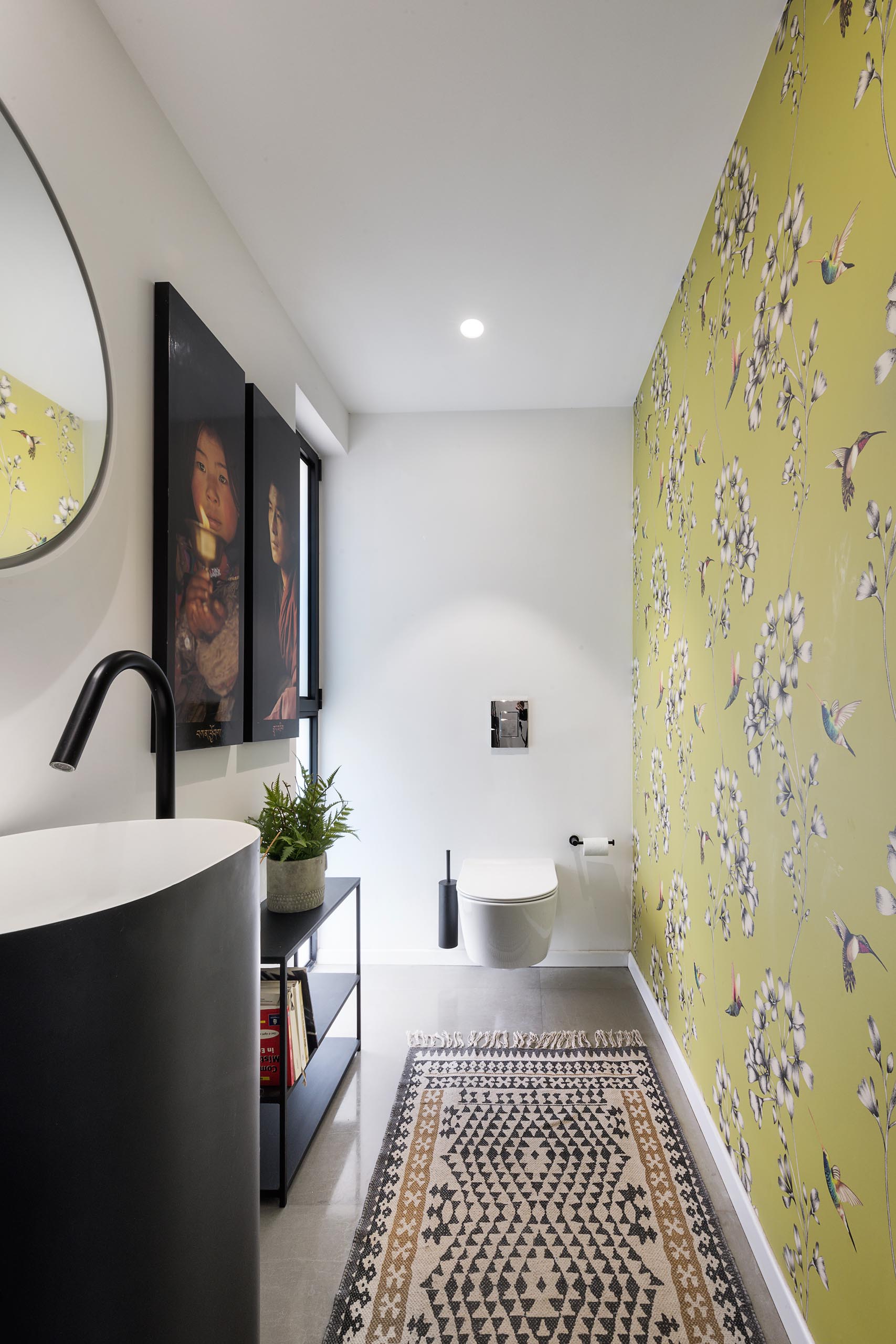 A powder room with a minimalist pedestal sink, black shelving, and a colorful yellow wallpaper with flowers and hummingbirds.