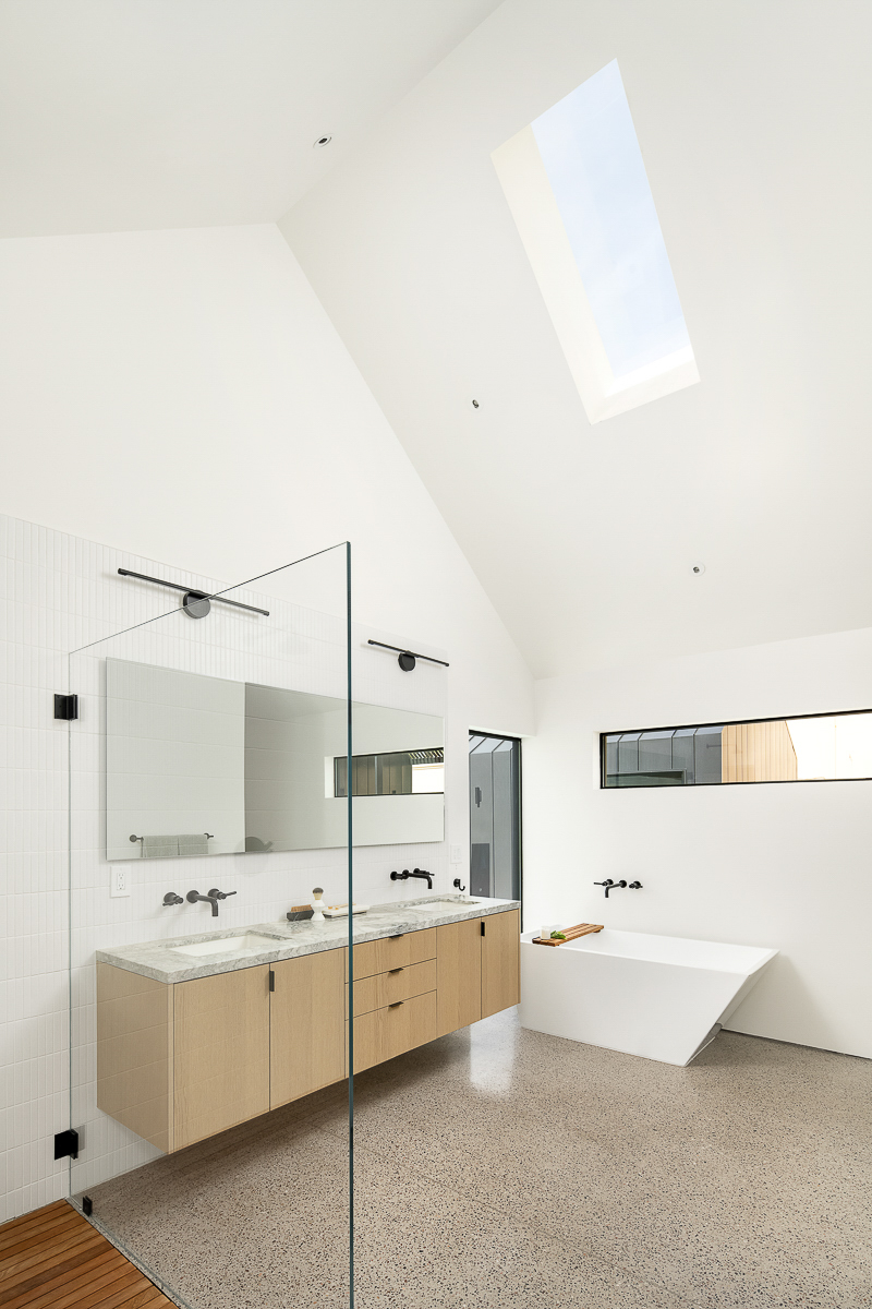 In this modern bathroom, the vaulted ceilings create a large and open space, while the white walls, ceiling, and skylight add brightness. In the shower white tiles complement the walls, while a wood floor in the shower adds a natural element to the space.