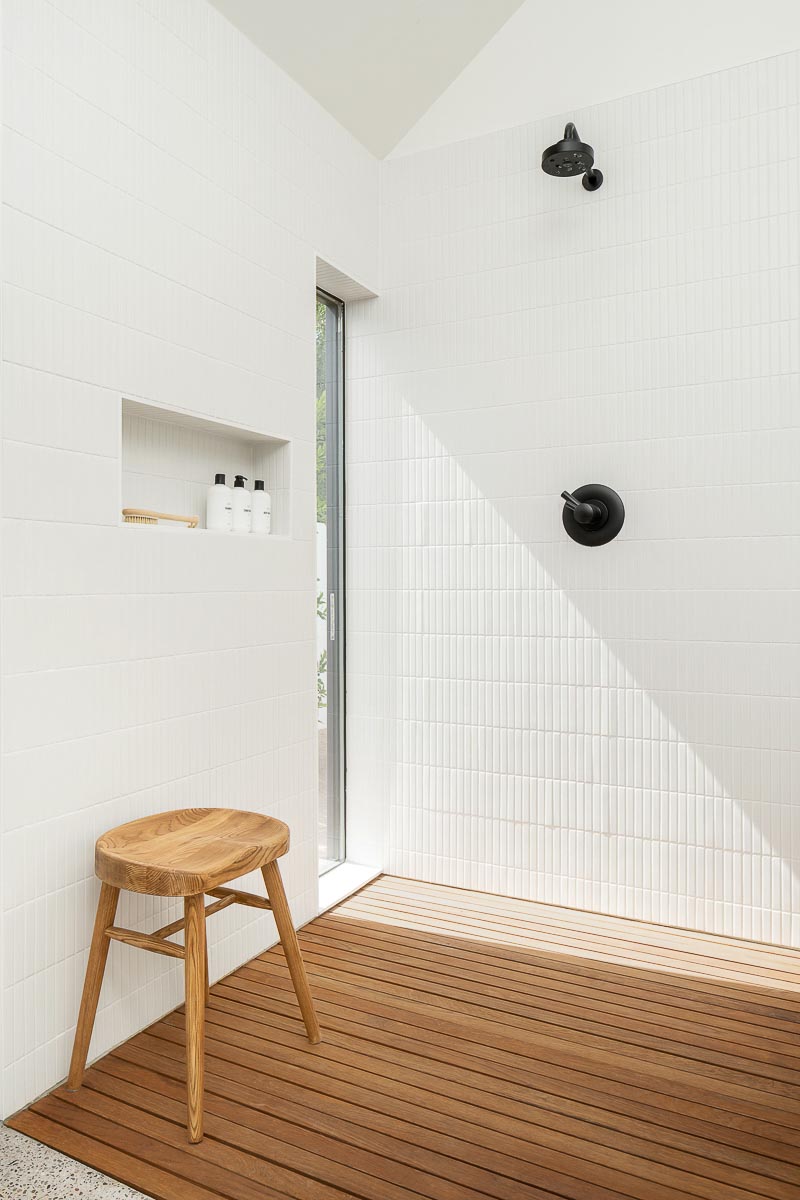 In this modern bathroom, the vaulted ceilings create a large and open space, while the white walls, ceiling, and skylight add brightness. In the shower white tiles complement the walls, while a wood floor in the shower adds a natural element to the space.