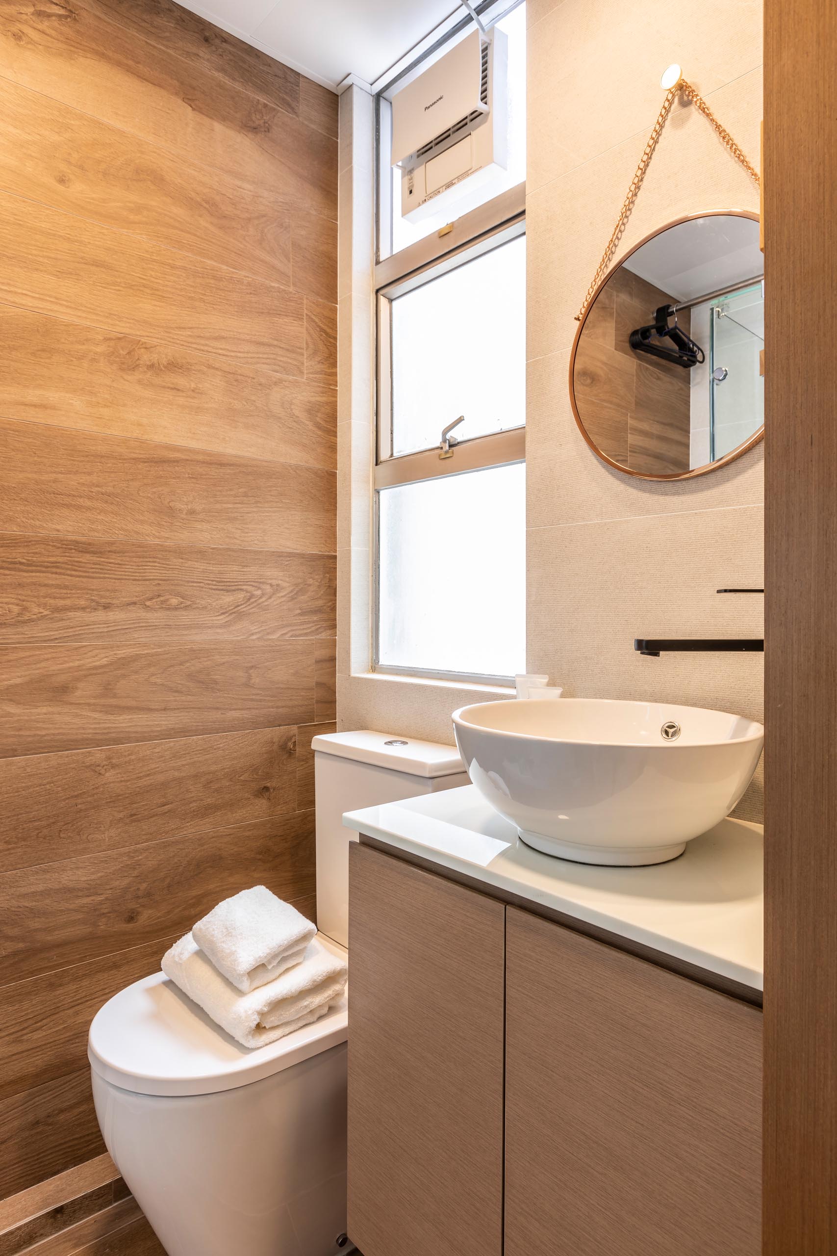 This modern bathroom in a small apartment includes a wood accent wall and a glass-enclosed shower.
