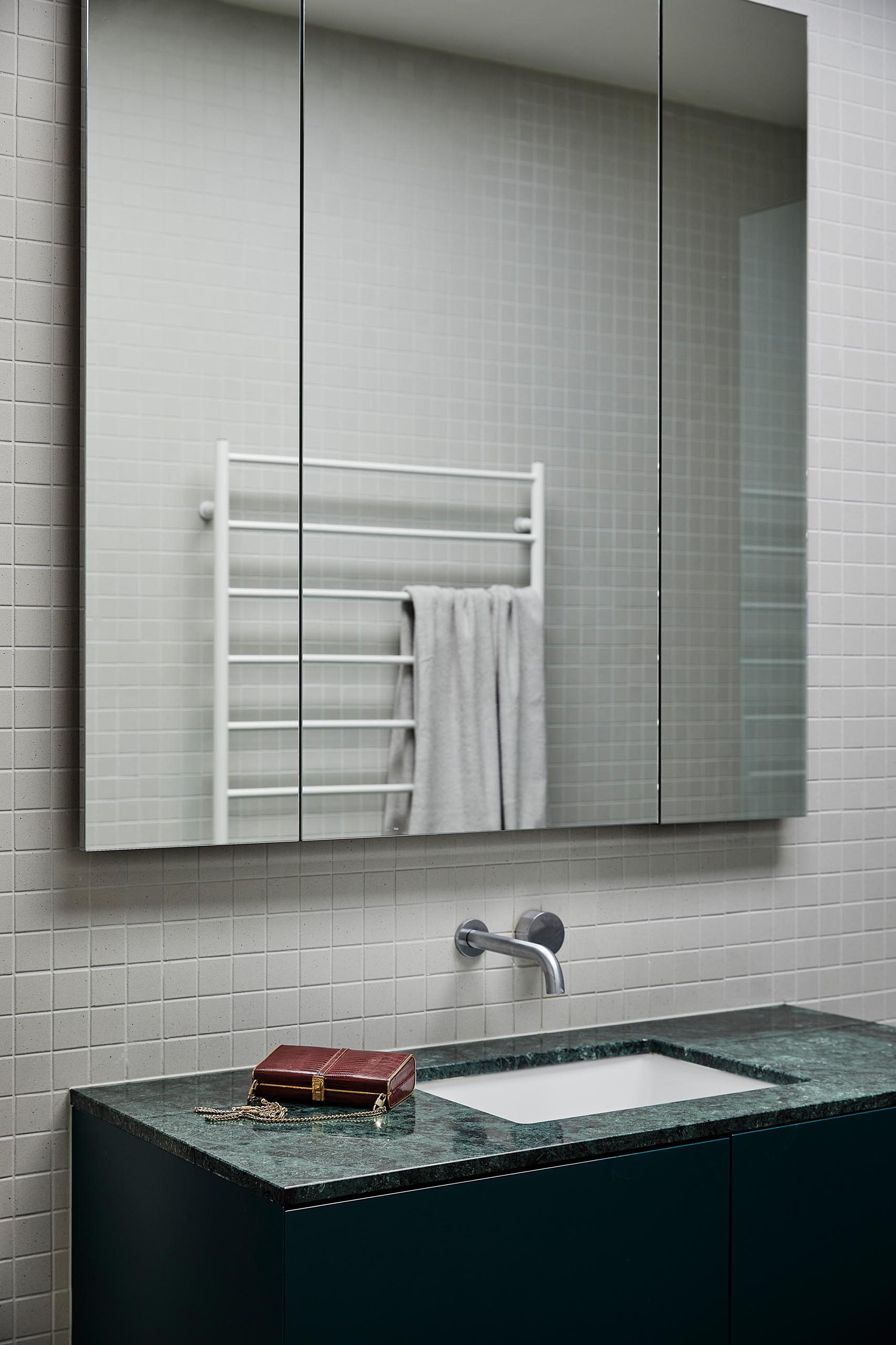 A modern bathroom with a a dark green vanity that contrasts the light gray square wall tiles and white basin.