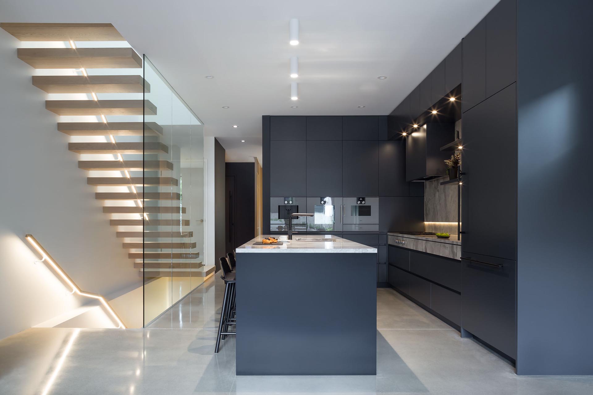 In this modern kitchen, there's black lacquered kitchen millwork, Gaggenau appliances and an integrated fridge, as well as marble countertops. Adjacent to the kitchen is a floating cantilevered wood staircase that includes a handrail with hidden lighting.