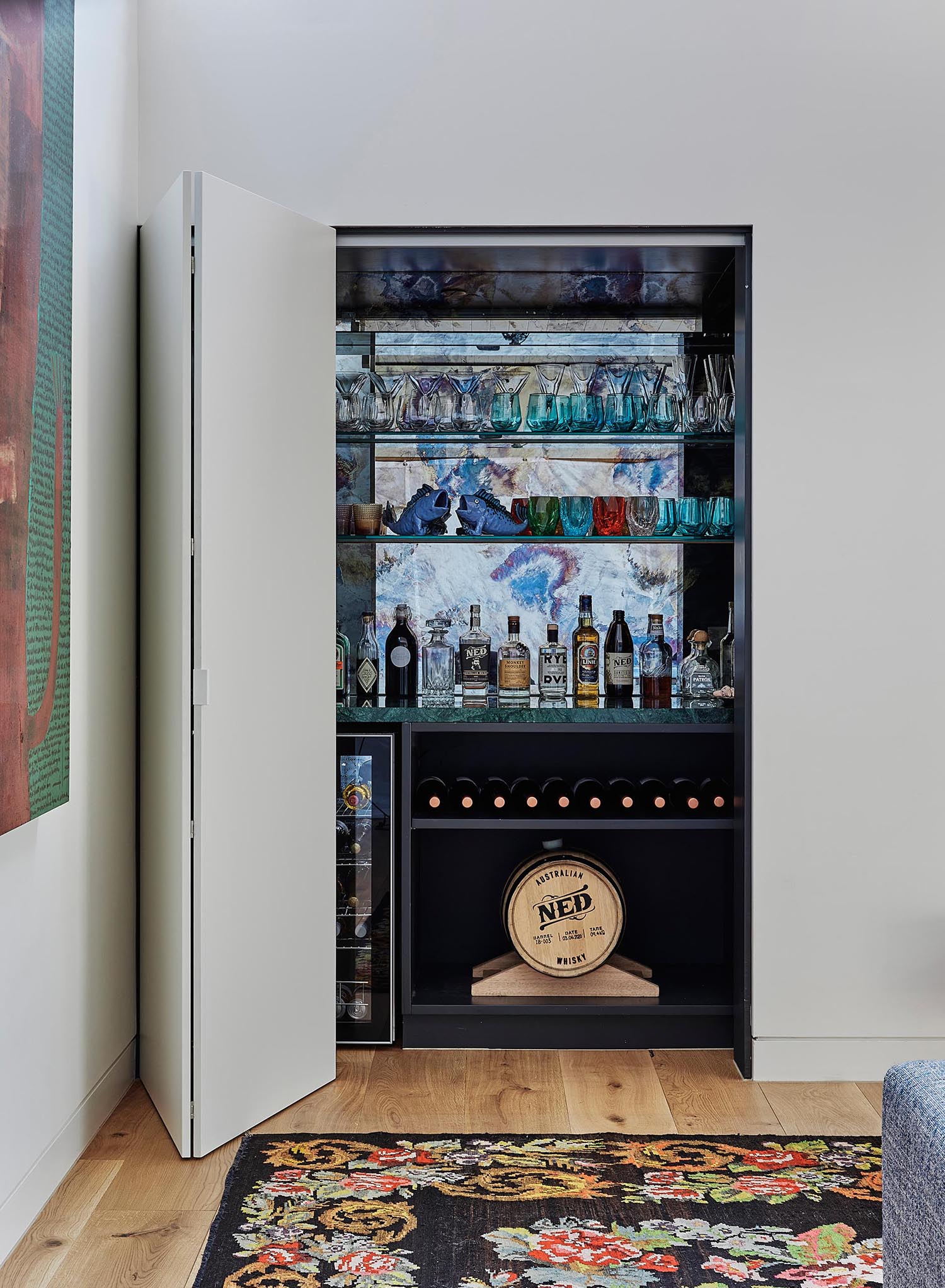 A home bar is hidden behind a closet in the living room, and includes a small fridge, bottle storage, and shelving for displaying glassware.