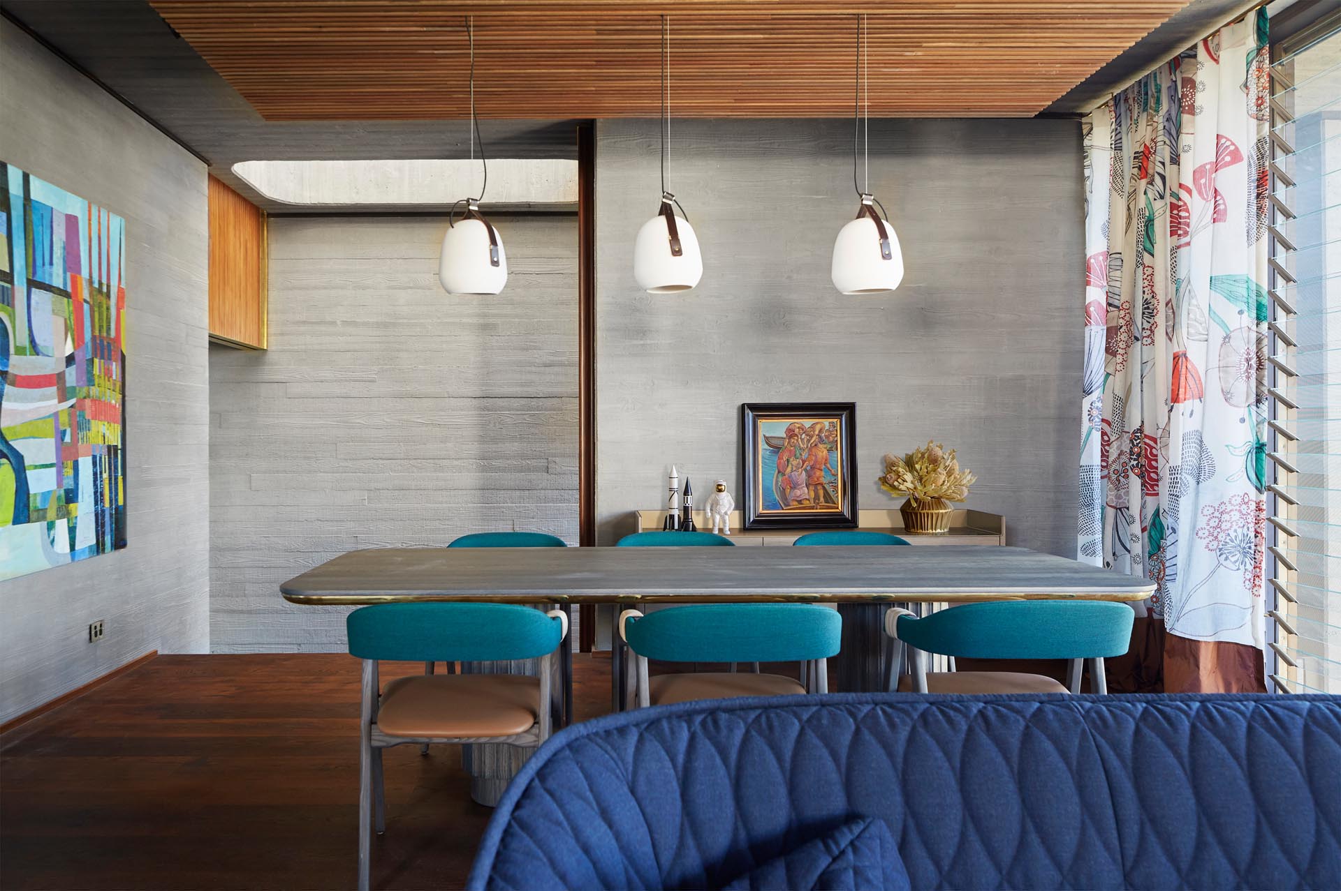 Textured concrete walls are visible in this modern dining room, that's been furnished with a wood table, dining chairs with blue accents, and three white pendant lamps.