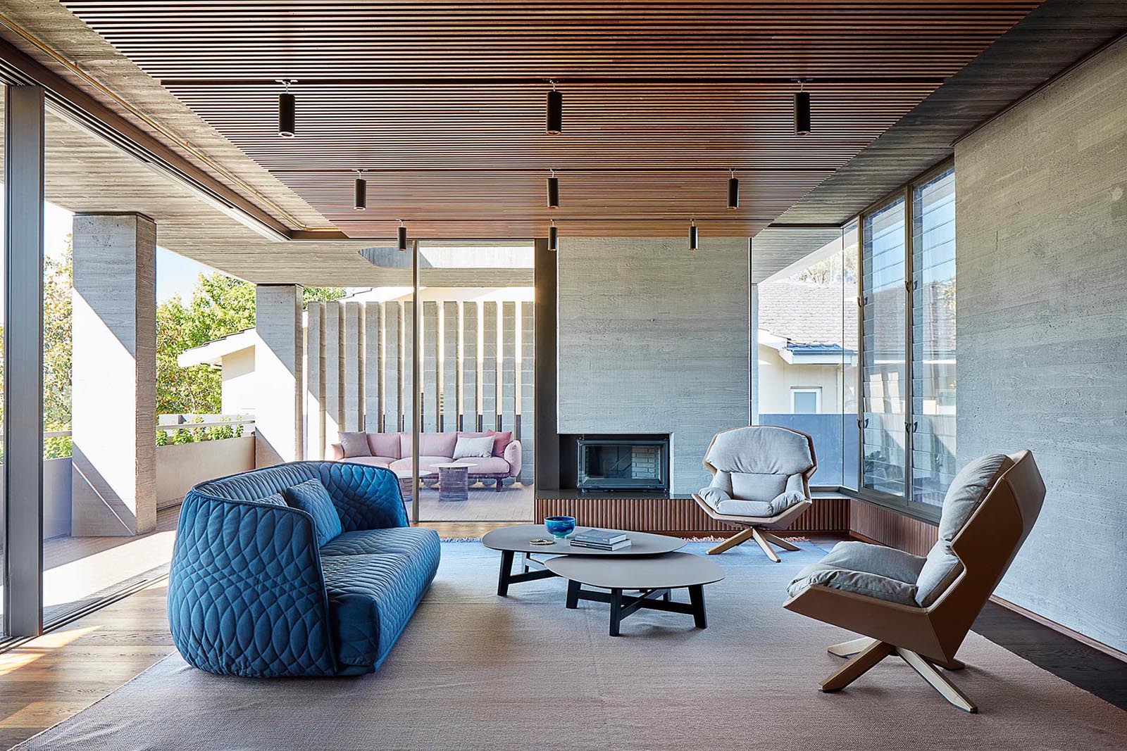 A modern living room with a fireplace that provides direct access to the covered outdoor area, and features a wood ceiling and board-formed concrete walls.