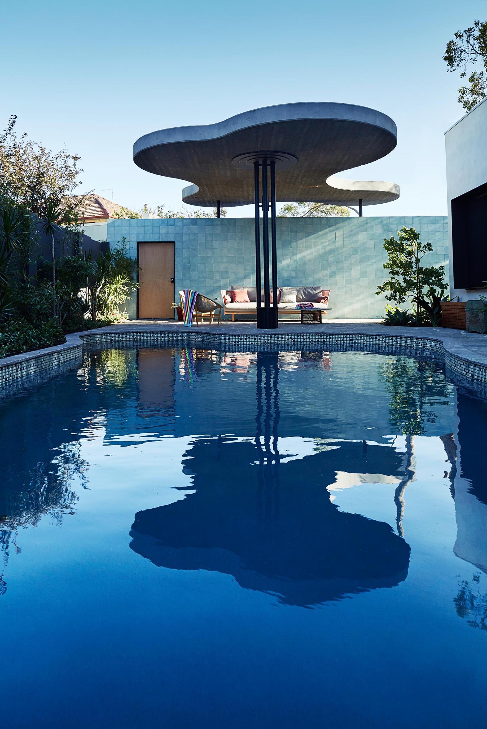 The organic shaped swimming pool of this modern home, is matched by the organic shape of the roof that provides shade to poolside furniture.