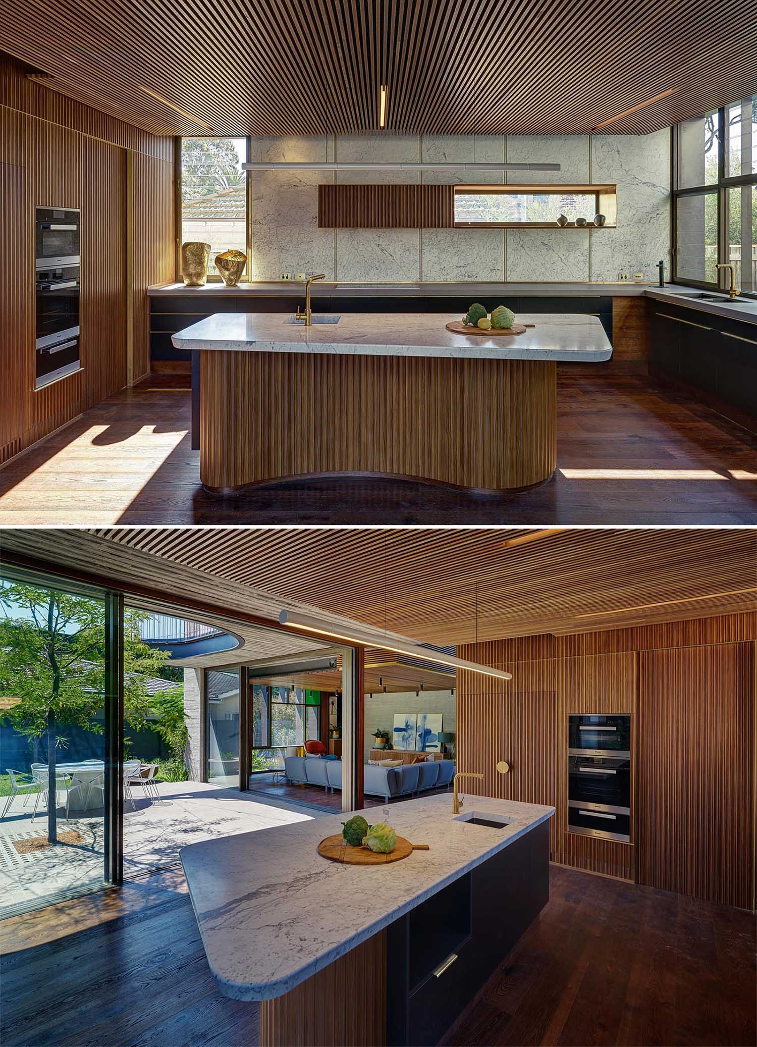 This modern kitchen has an island with a solid marble slab countertop and organically shaped timber cabinetwork. Timber ceilings conceal acoustic insulation.