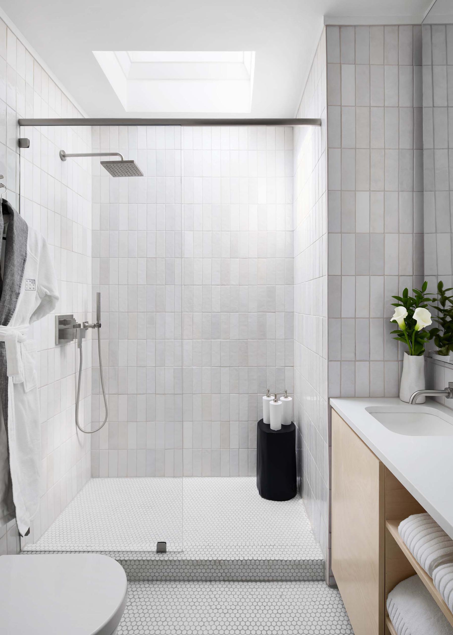 In this modern spa-inspired bathroom, penny tiles cover the floor, a wood vanity with open shelving is topped with a white countertop, and an oversized skylight provides natural light for the tiled shower.