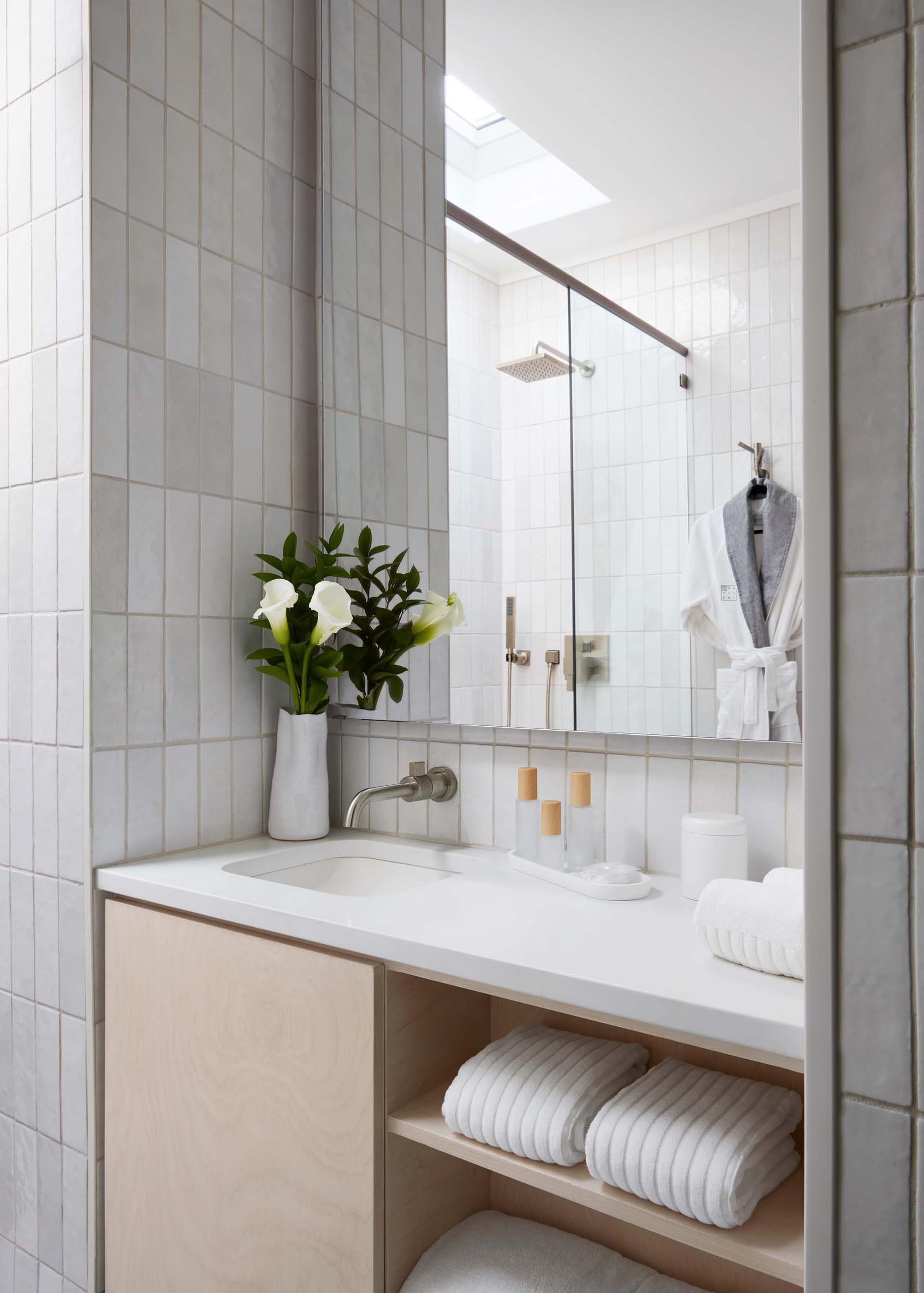 This modern bathroom has been designed with penny tiles covering the floor, a wood vanity with open shelving that's topped with a white countertop, and an oversized skylight that provides natural light for the tiled shower.