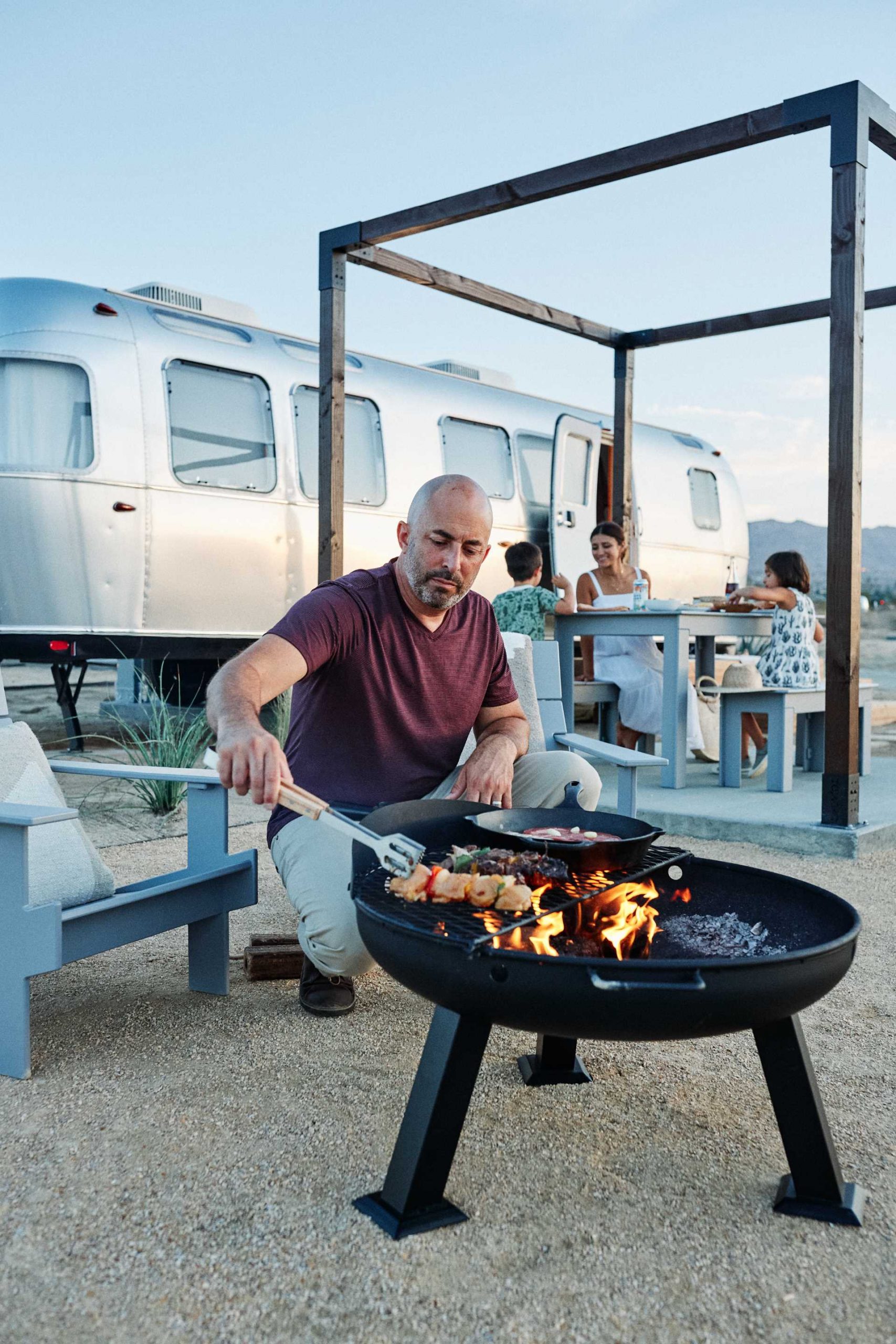 An Airstream located in the desert includes an outdoor dining area, a small seating area with a firepit, and wood stairs.