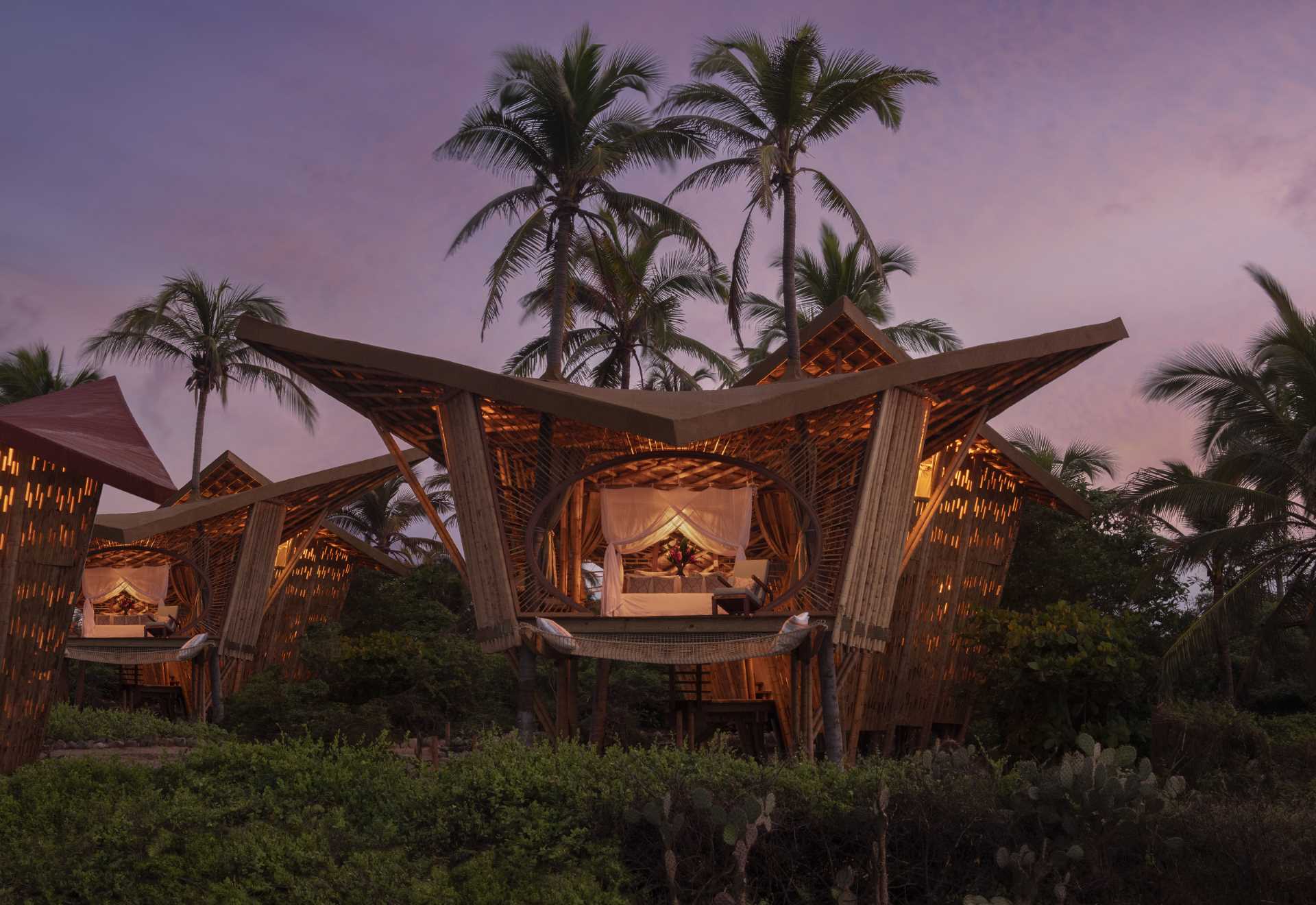 At night, the lighting within these treehouses creates a warm glow, highlighting the bamboo structure.