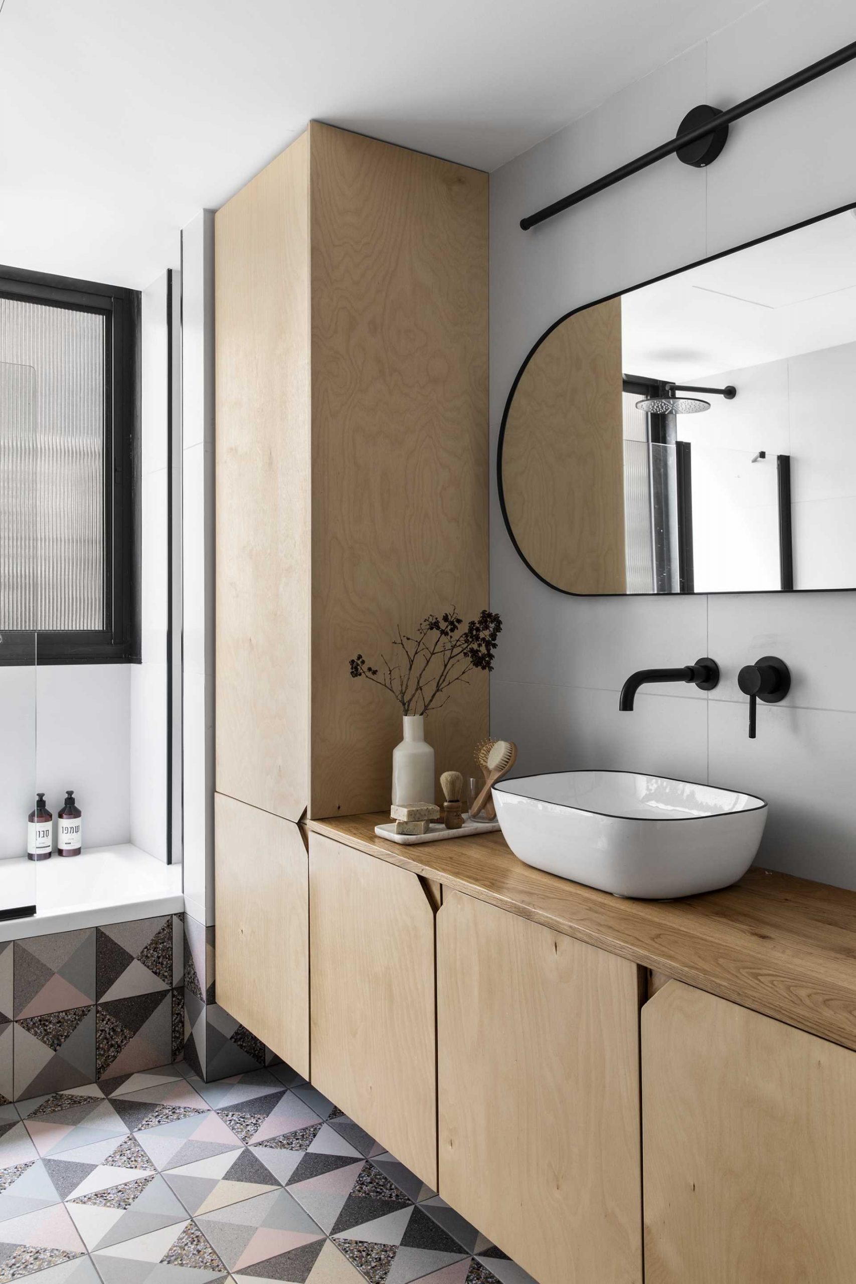 This modern bathroom is spacious and bright, and includes a birch wood vanity and cabinet with unique triangular CNC handles. Geometric terrazzo tiles add a touch of color to the space.