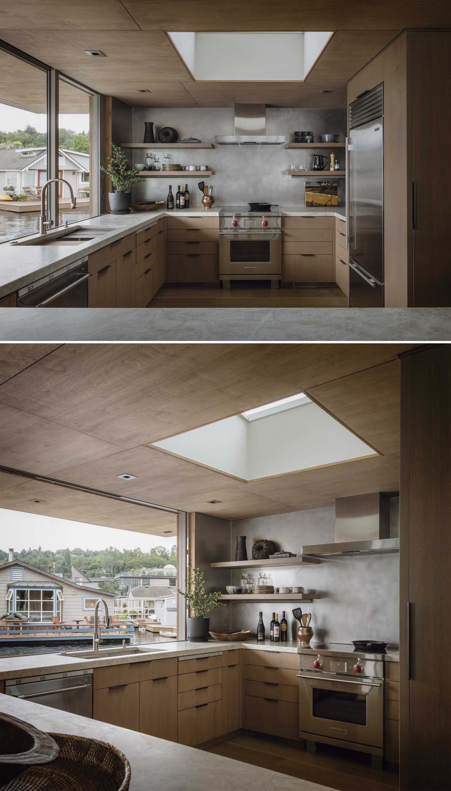 This modern kitchen is slightly raised from the nearby dining area and includes open shelving, a skylight, wood cabinetry, and large windows.