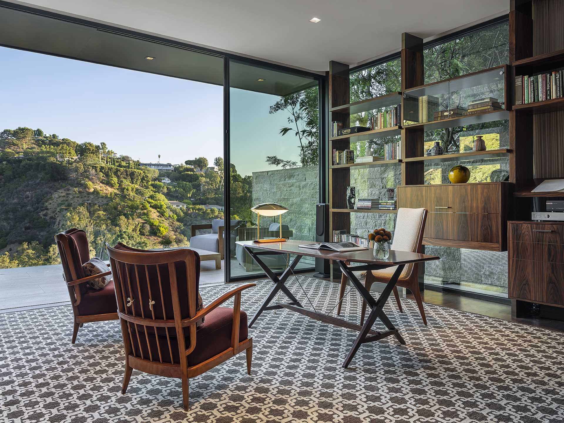 A modern home office with glass walls, access to a patio, and built-in wood shelving.