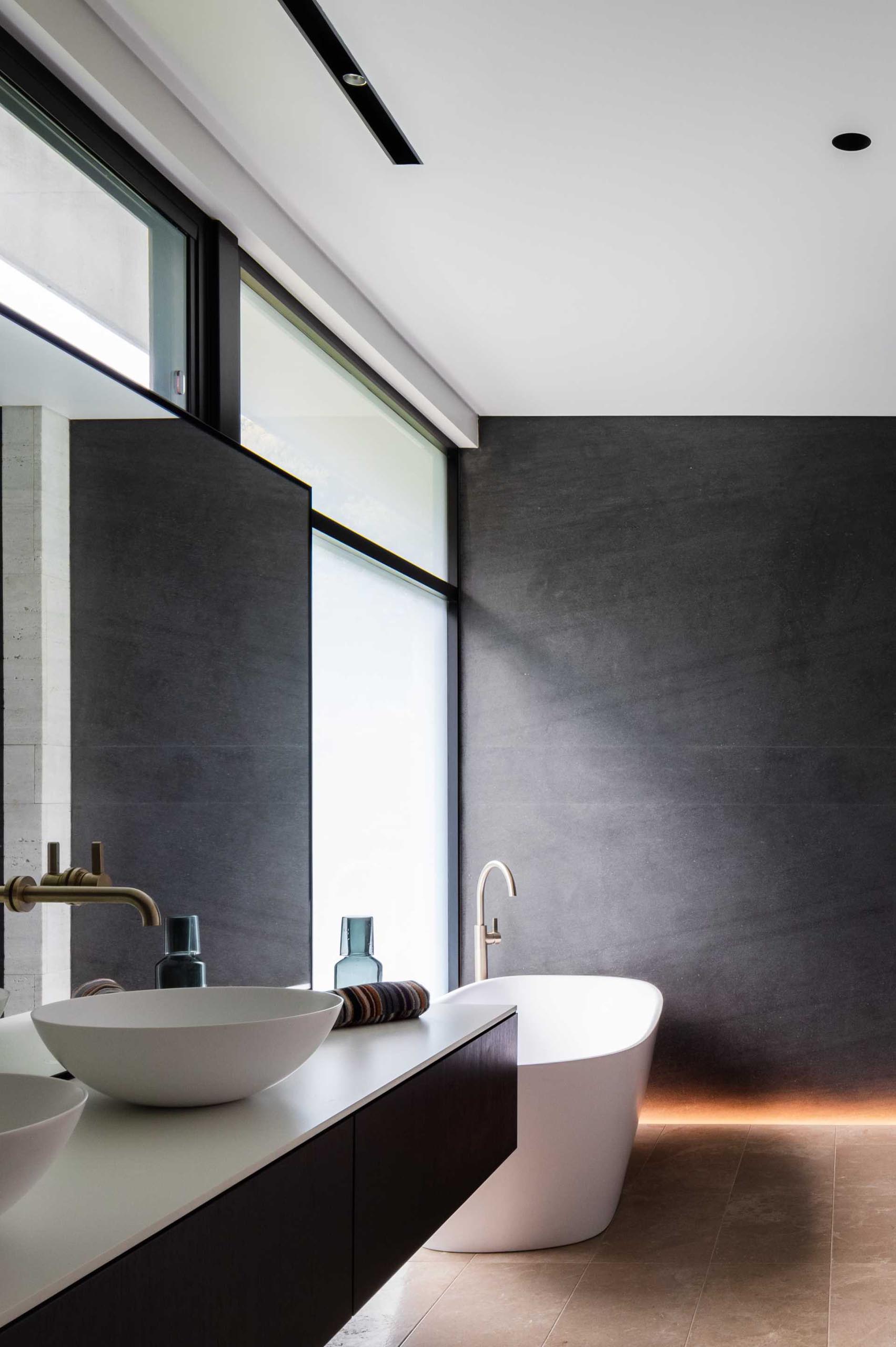 In this modern bathroom, the black window frames and vanity complement the dark accent wall, while a freestanding bathtub has been placed in front of a window that overlooks the backyard.