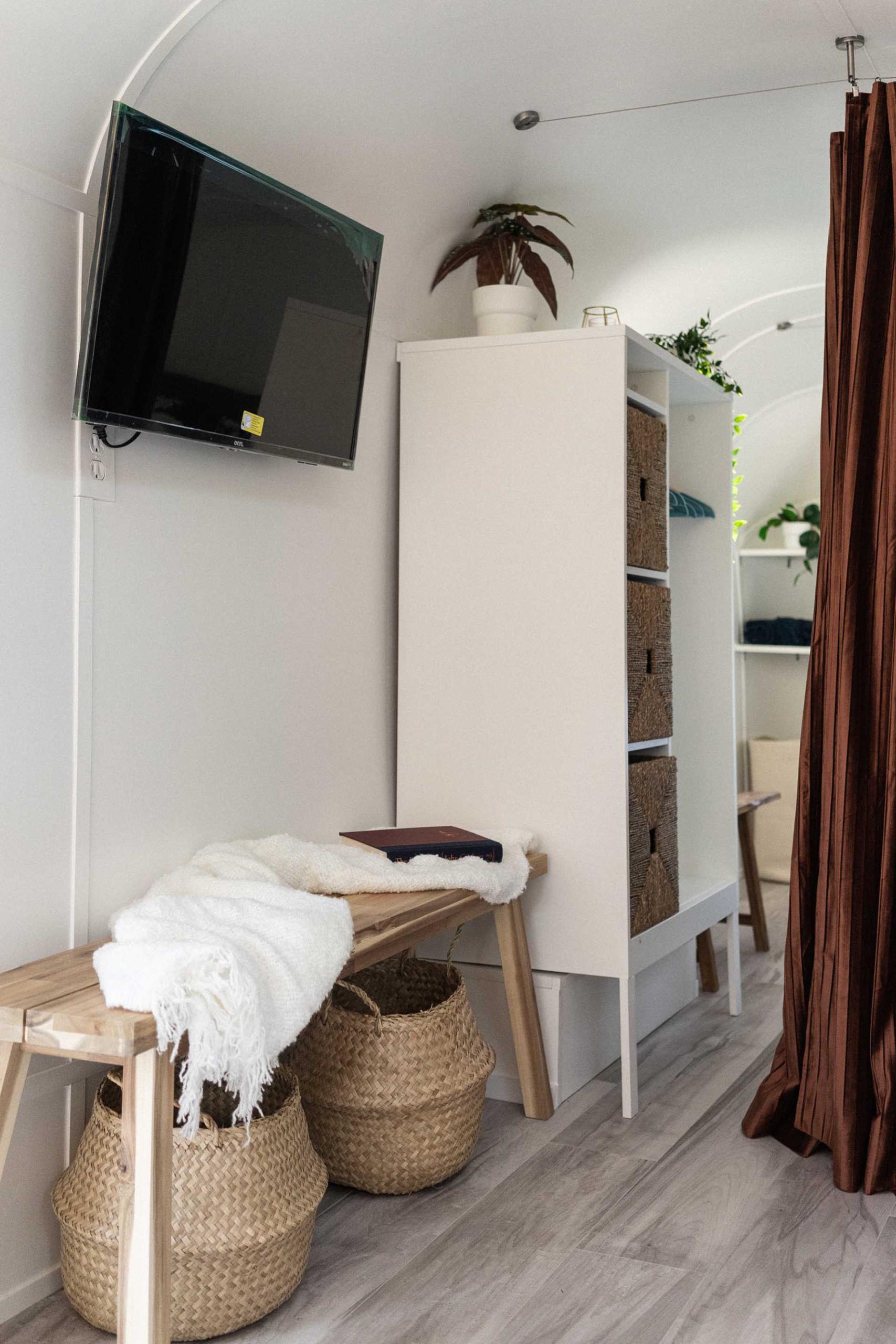 The entryway of this renovated vintage travel trailer includes a bench with a couple of storage baskets, and a wall-mounted tv.