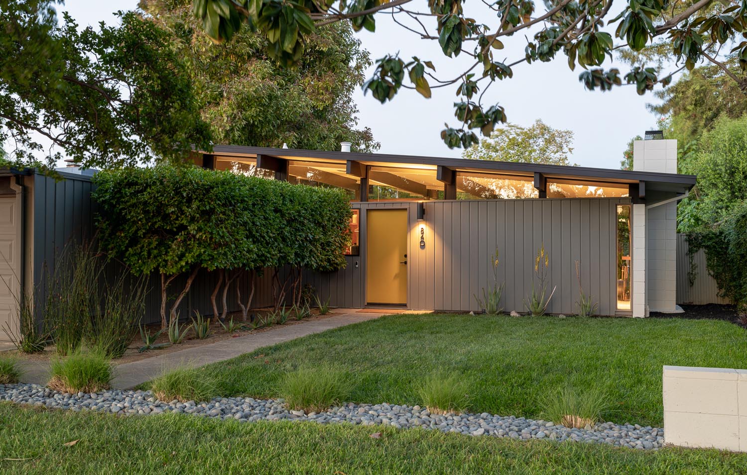 The new facade of this Eichler house has a light brown exterior which allows the plants to be showcased, while the concrete patio was replaced with grass.