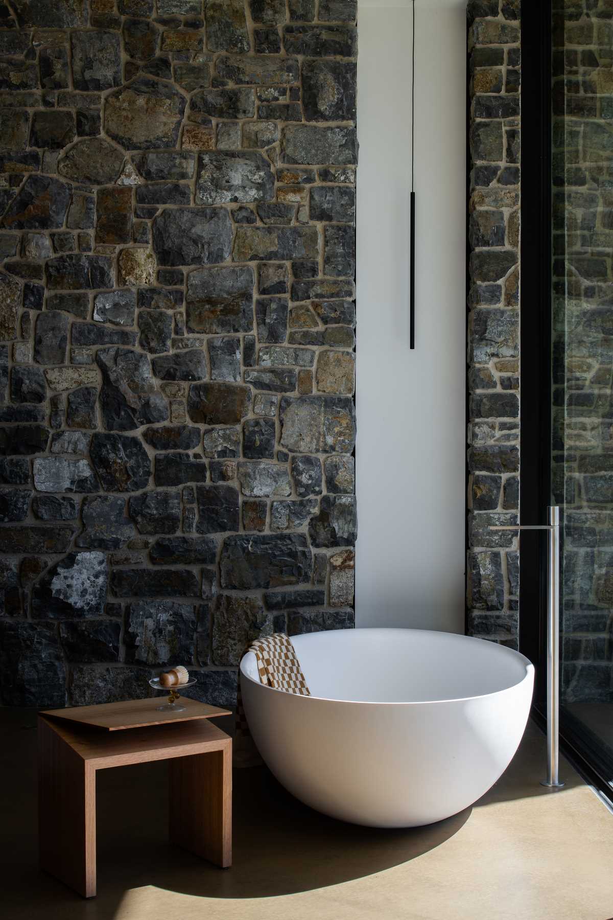 In this modern en-suite bathroom, there's stone walls, and a freestanding white bathtub positioned in front of the windows to take advantage of the ocean views