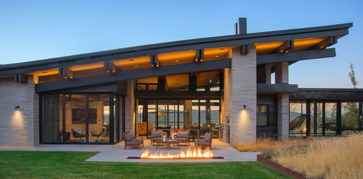 This modern house includes a outdoor dining and BBQ area that's covered by the sloping roof. Nearby there's a sitting area, a fireplace, and yard.