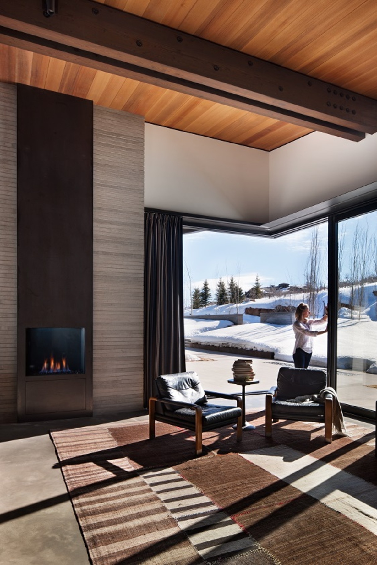 In this modern room, a fireplace with a black surround draws the eye up to the high ceilings and exposed wood beams. Sliding glass doors open the interior to the outdoors.