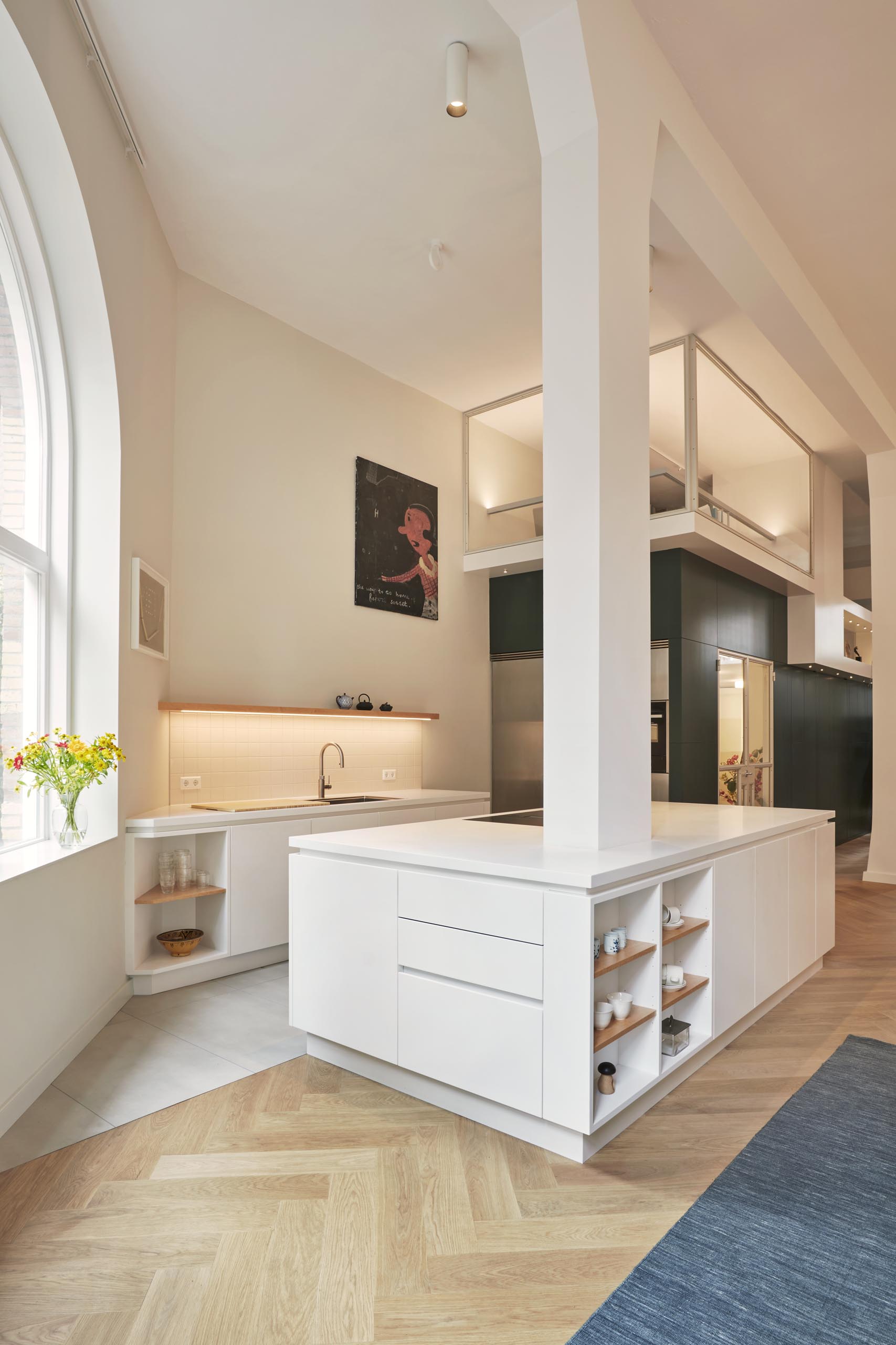 A modern white kitchen with a large island that wraps around a column, and includes both drawers and open shelving.