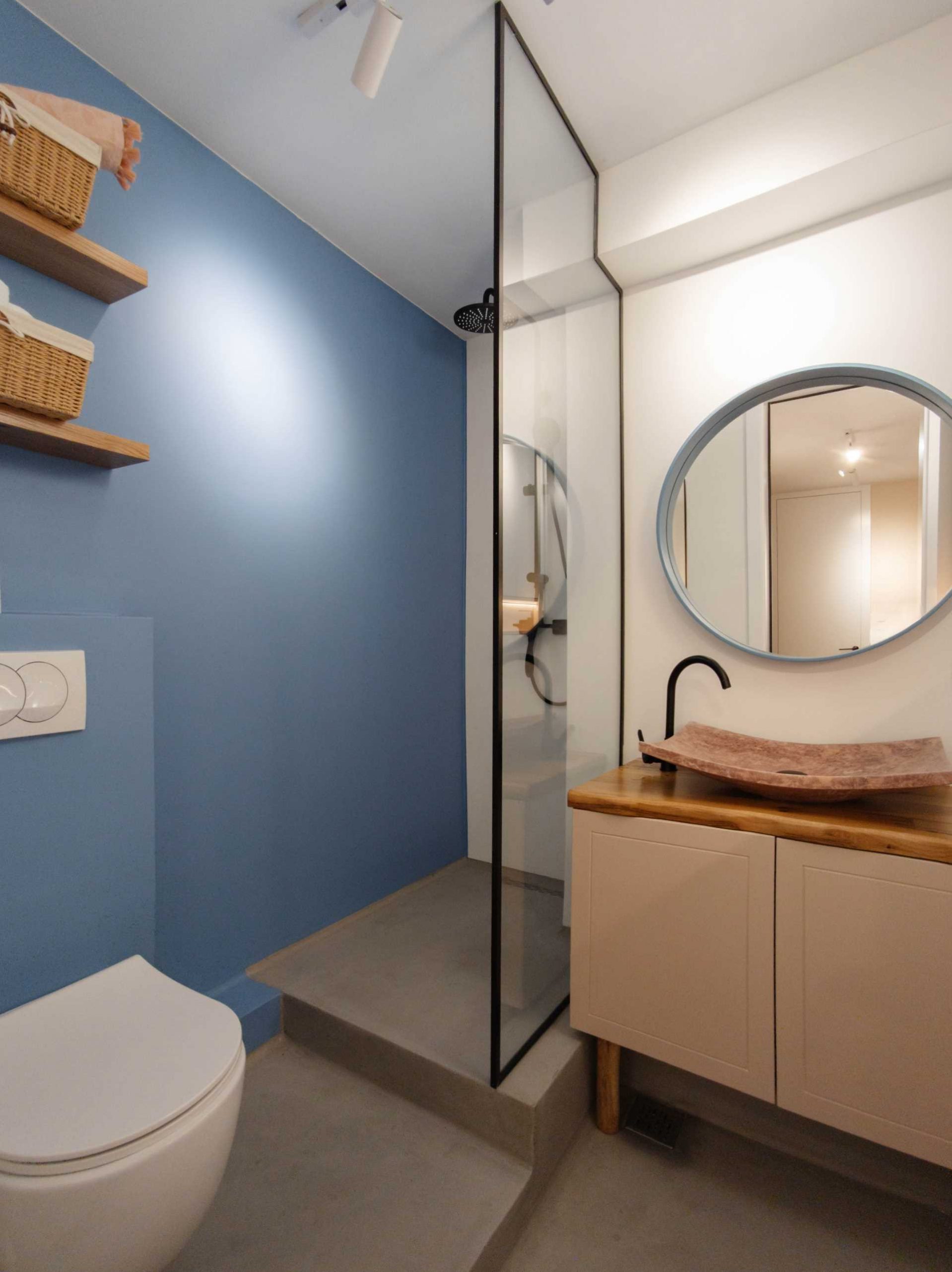In this small and contemporary bathroom, the shower has a matte blue wall and a black framed glass shower screen. There's also floating wood shelves, a wood vanity with a blue-framed round mirror above, and a built-in cabinet with a shelving niche.