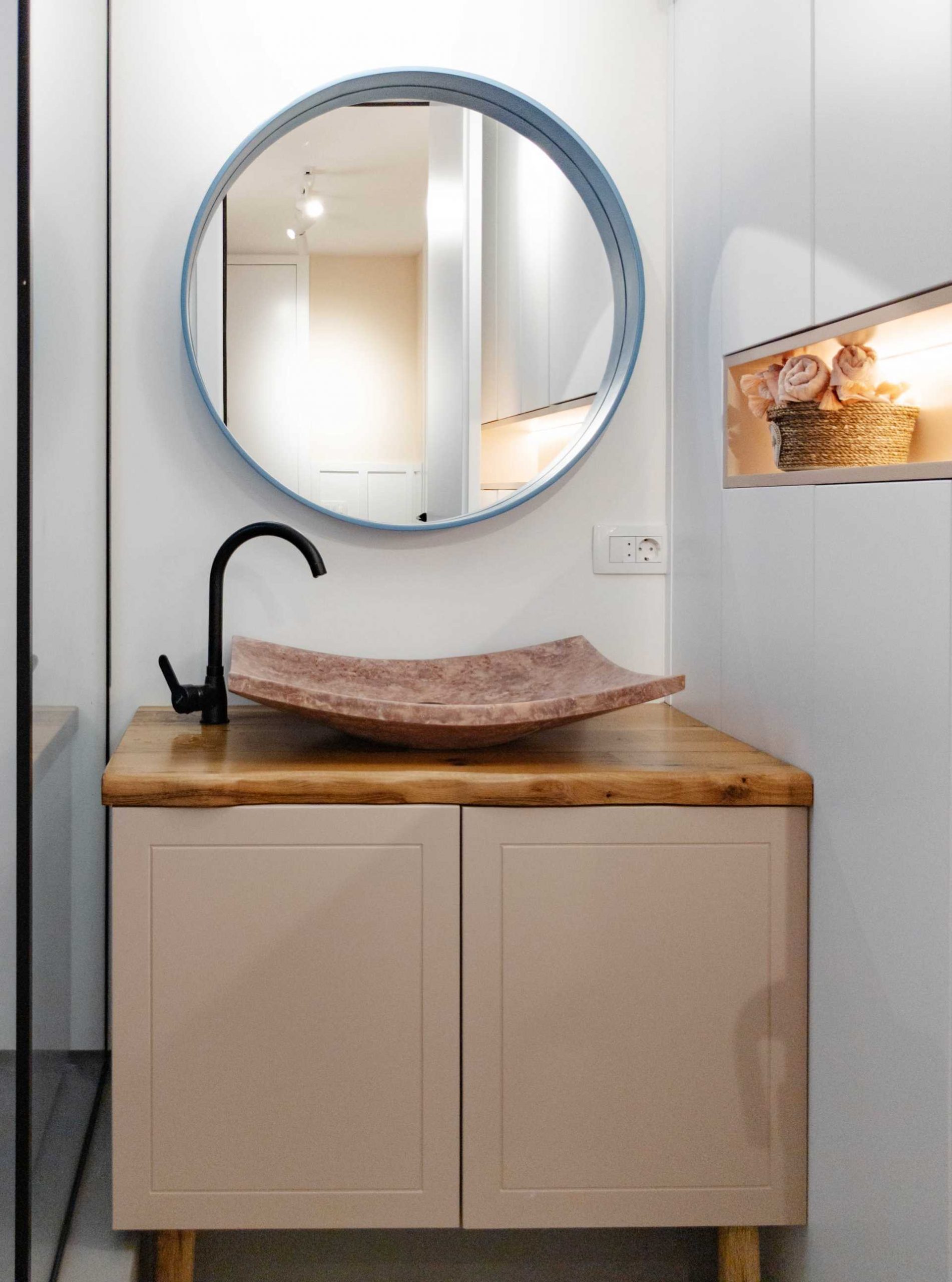 In this small and contemporary bathroom, there's a wood vanity with a blue-framed round mirror above, and a built-in cabinet with a shelving niche.