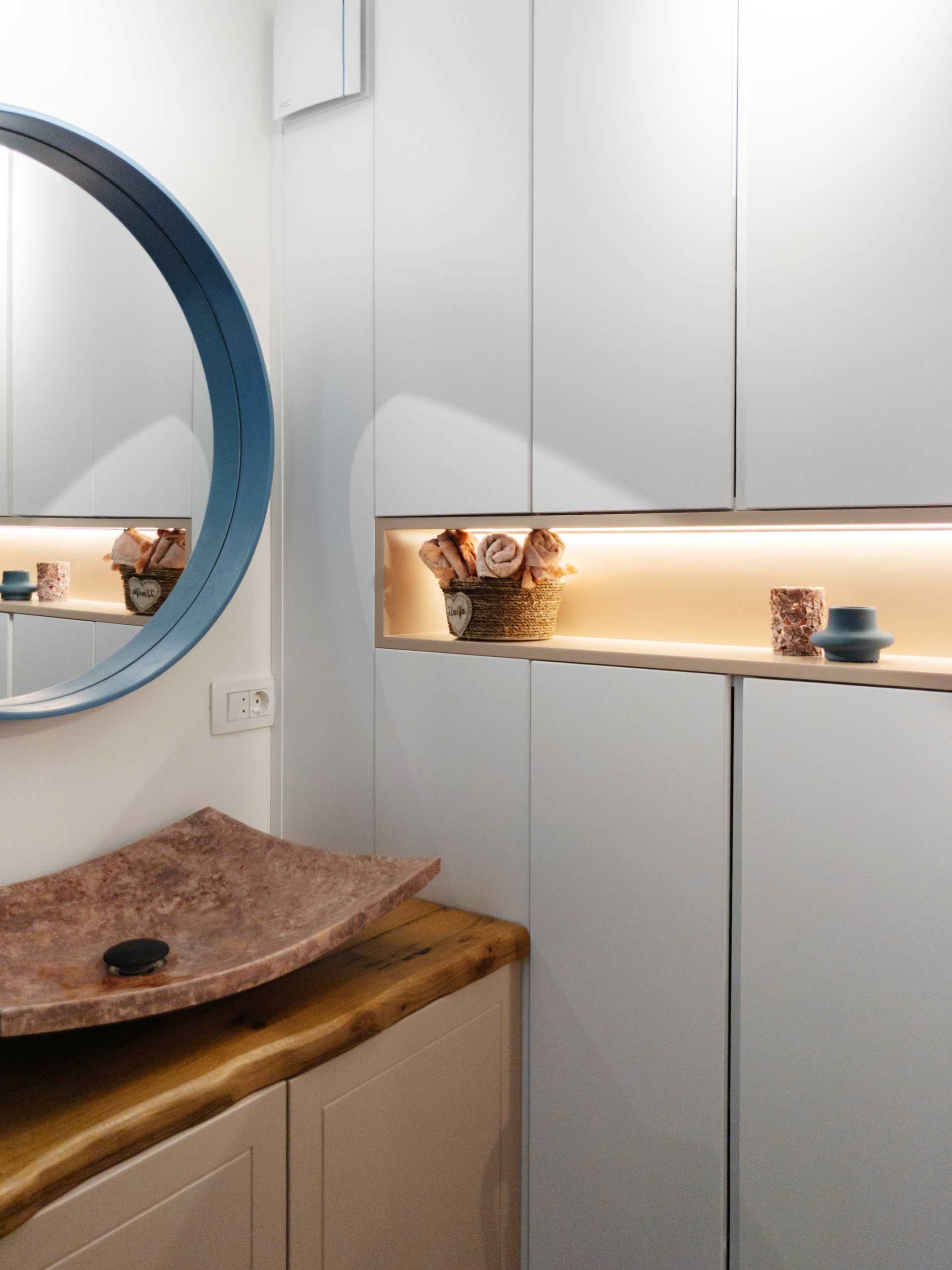 In this small and contemporary bathroom, there's a wood vanity with a blue-framed round mirror above, and a built-in cabinet with a shelving niche.