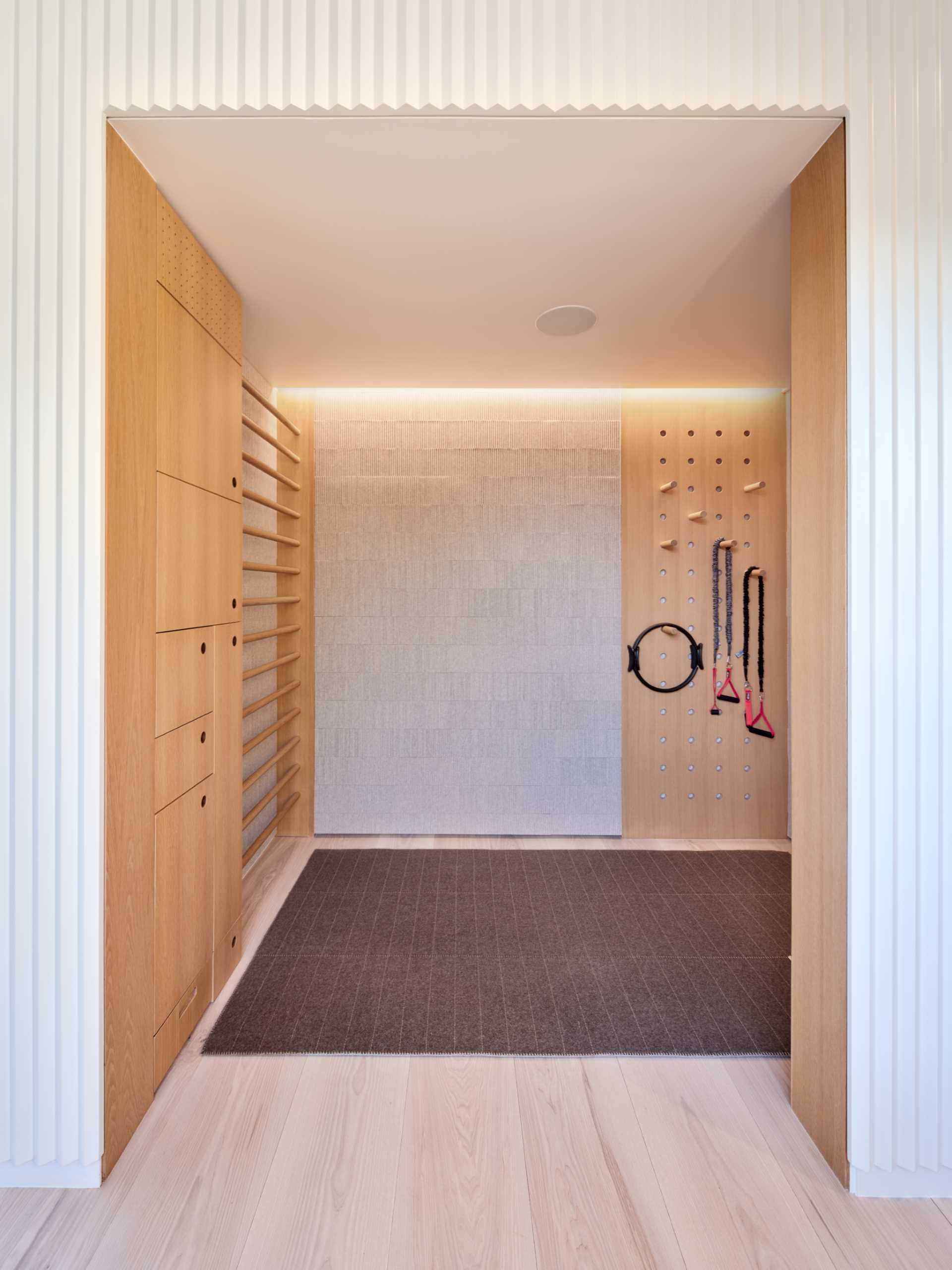 A wellness room for stretching and meditation, that's lined with softly ridged millwork that conceals storage cabinet to accommodate yoga equipment, while white oak and felt wall panels and a custom ladder/peg system complete the space.