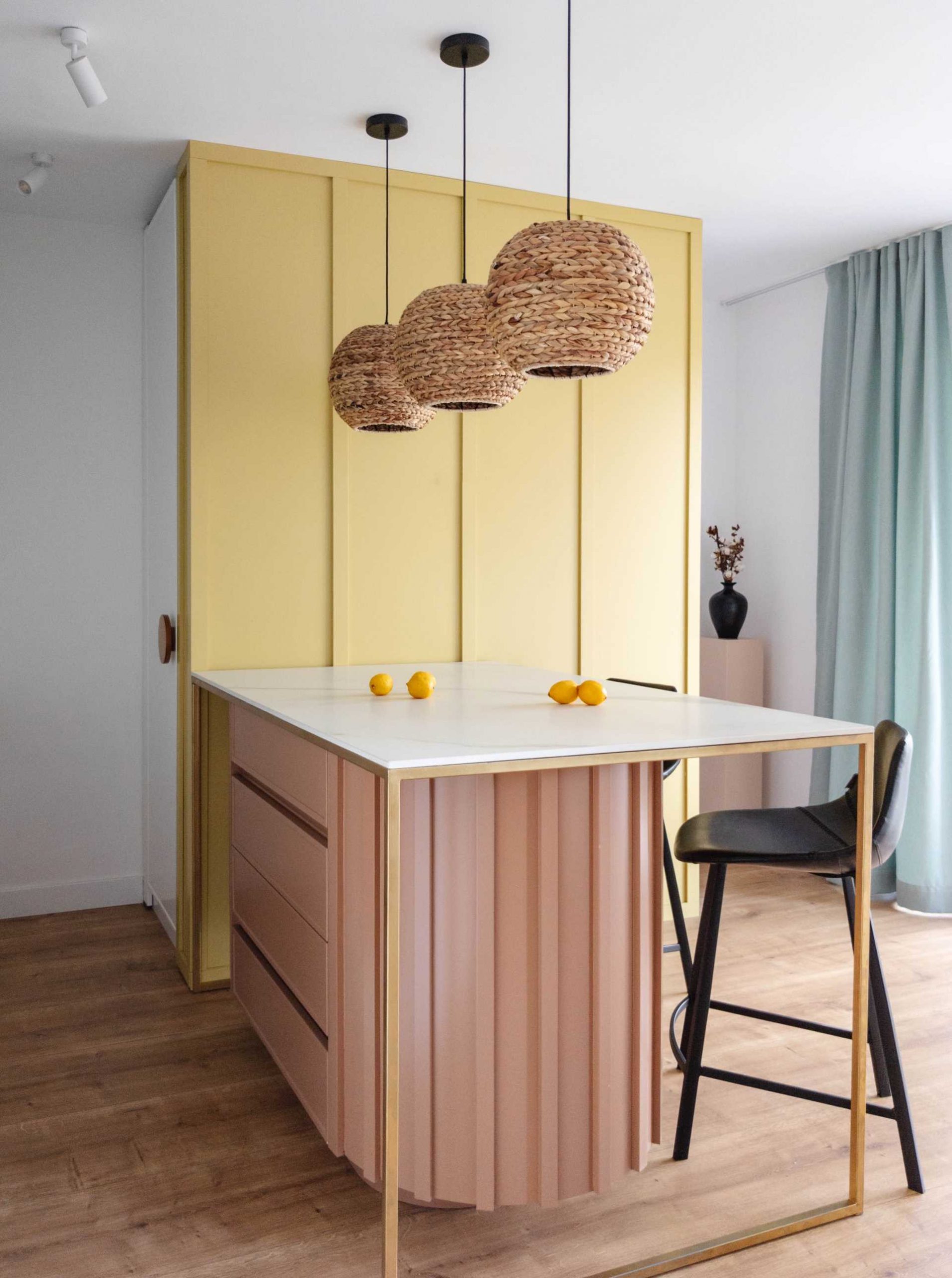 Pastel colors have been used to brighten up this contemporary kitchen, with a yellow accent wall, a curved light pink base for the peninsula, and woven pendant lights that add a natural touch to the space.