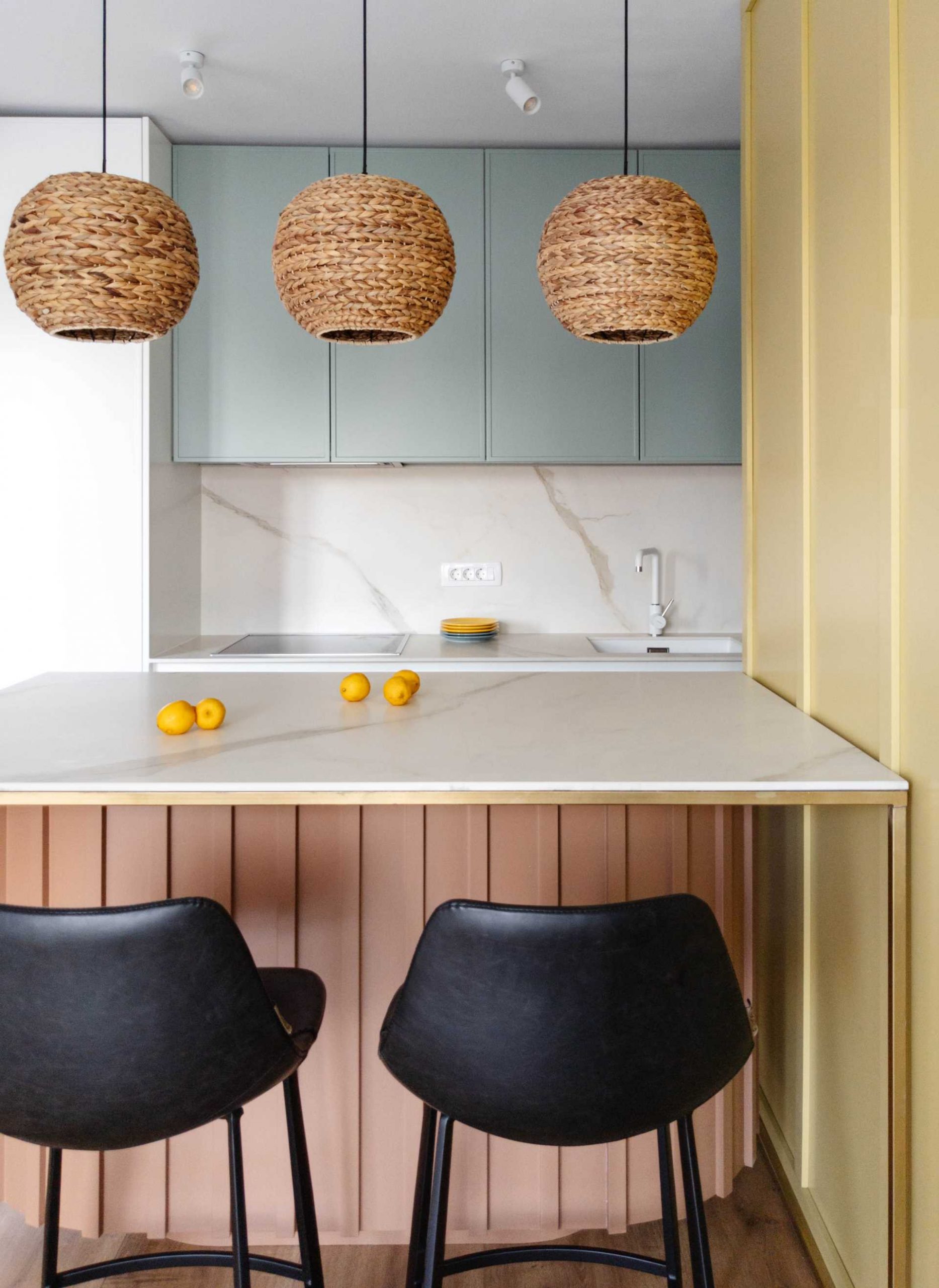 Pastel colors have been used to brighten up this contemporary kitchen, with a yellow accent wall, a curved light pink base for the peninsula, and muted mint green for the upper cabinets. Woven pendant lights add a natural touch to the space.