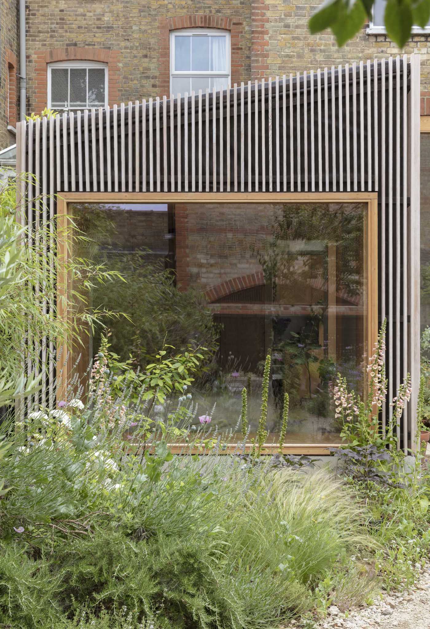 A modern home addition with a large picture window, is clad in wood slats.