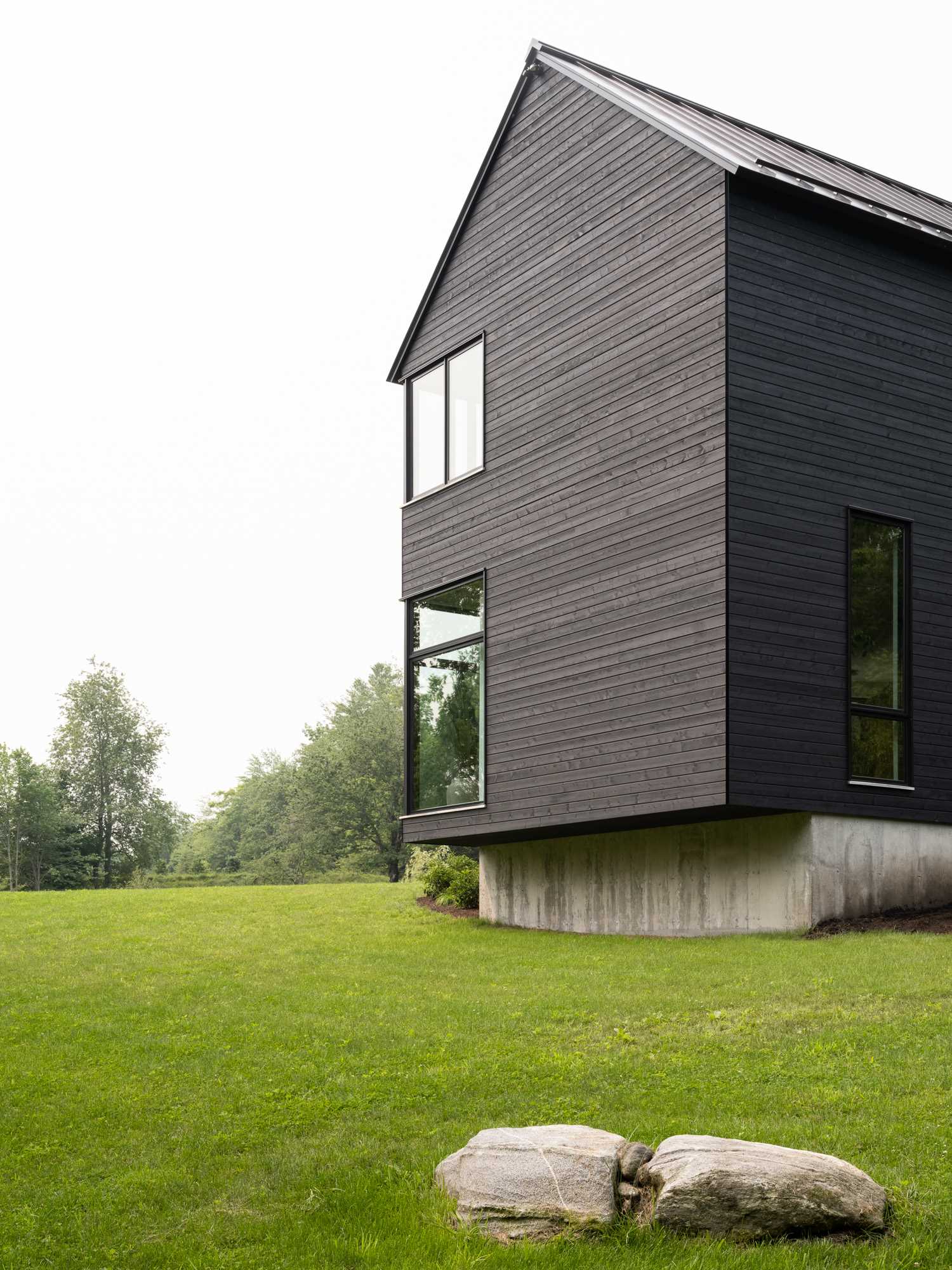 This modern house, surrounded by trees and grass, has a bold exterior clad with black stained wood siding and a standing seam metal roof.