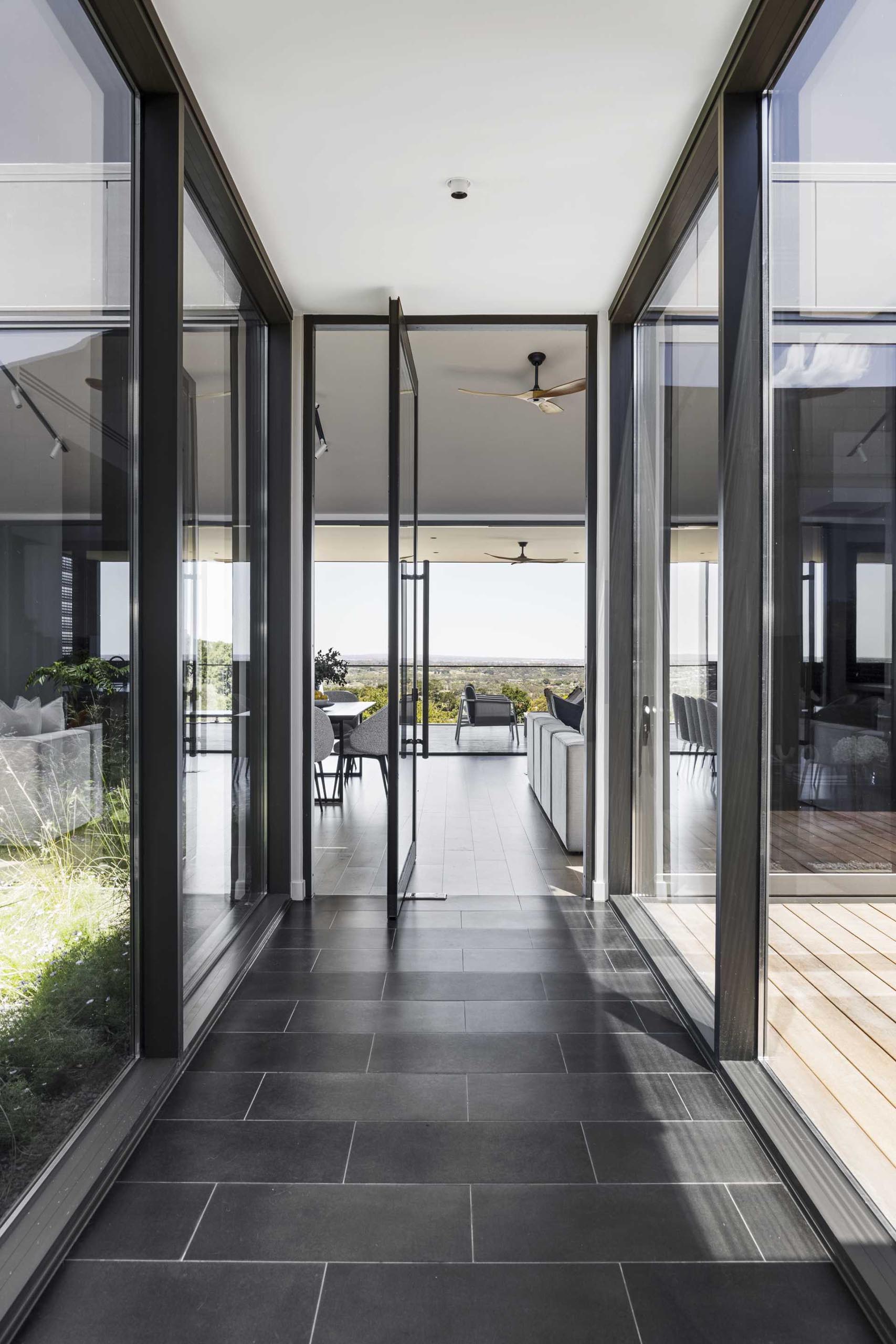 A modern corridor opens up to a glass-lined walkway where a pivoting glass door welcomes people to the open plan social areas.