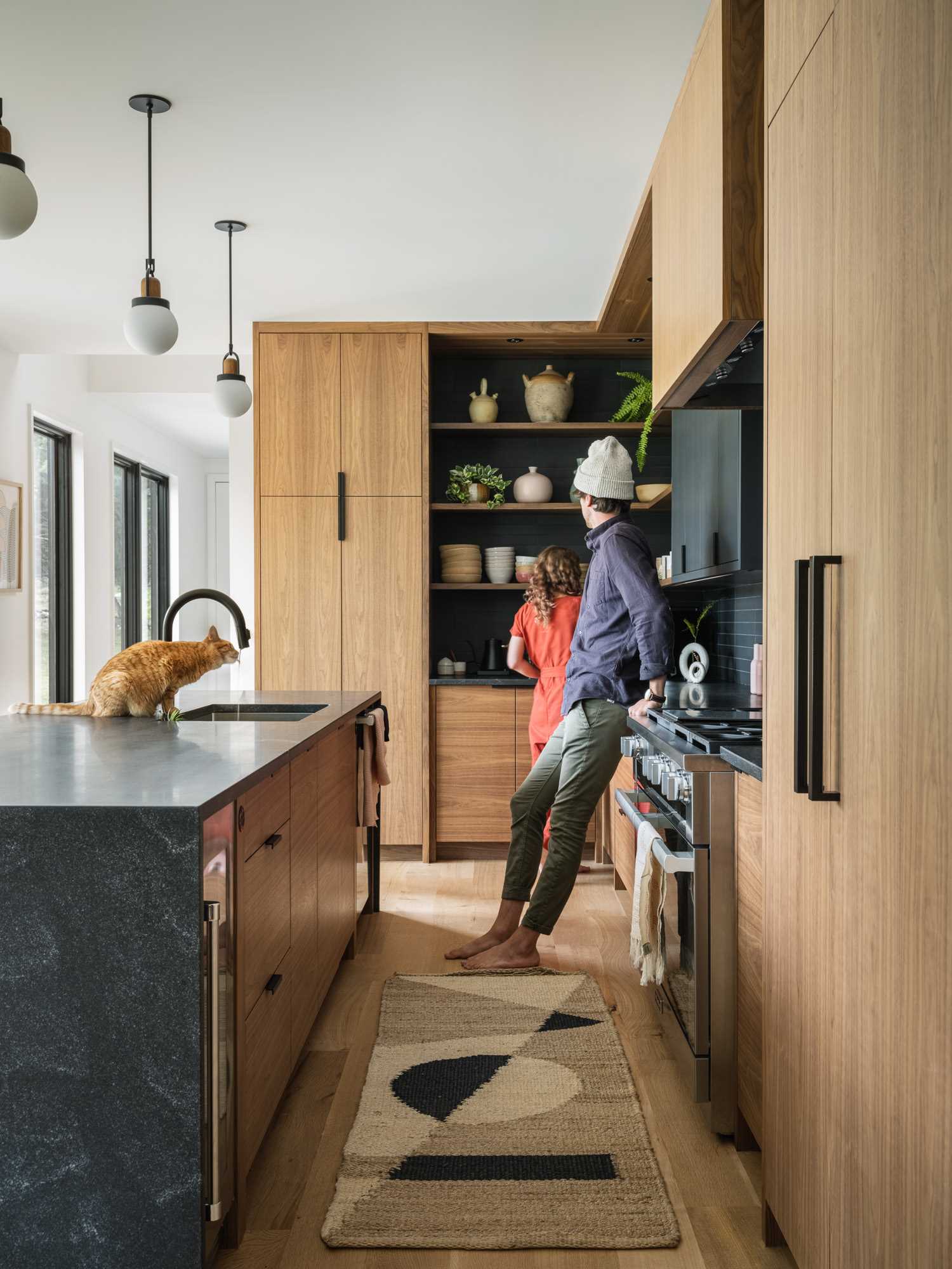 A modern kitchen with an island, includes wood cabinets, open corner shelves, and black rectangular tiles.