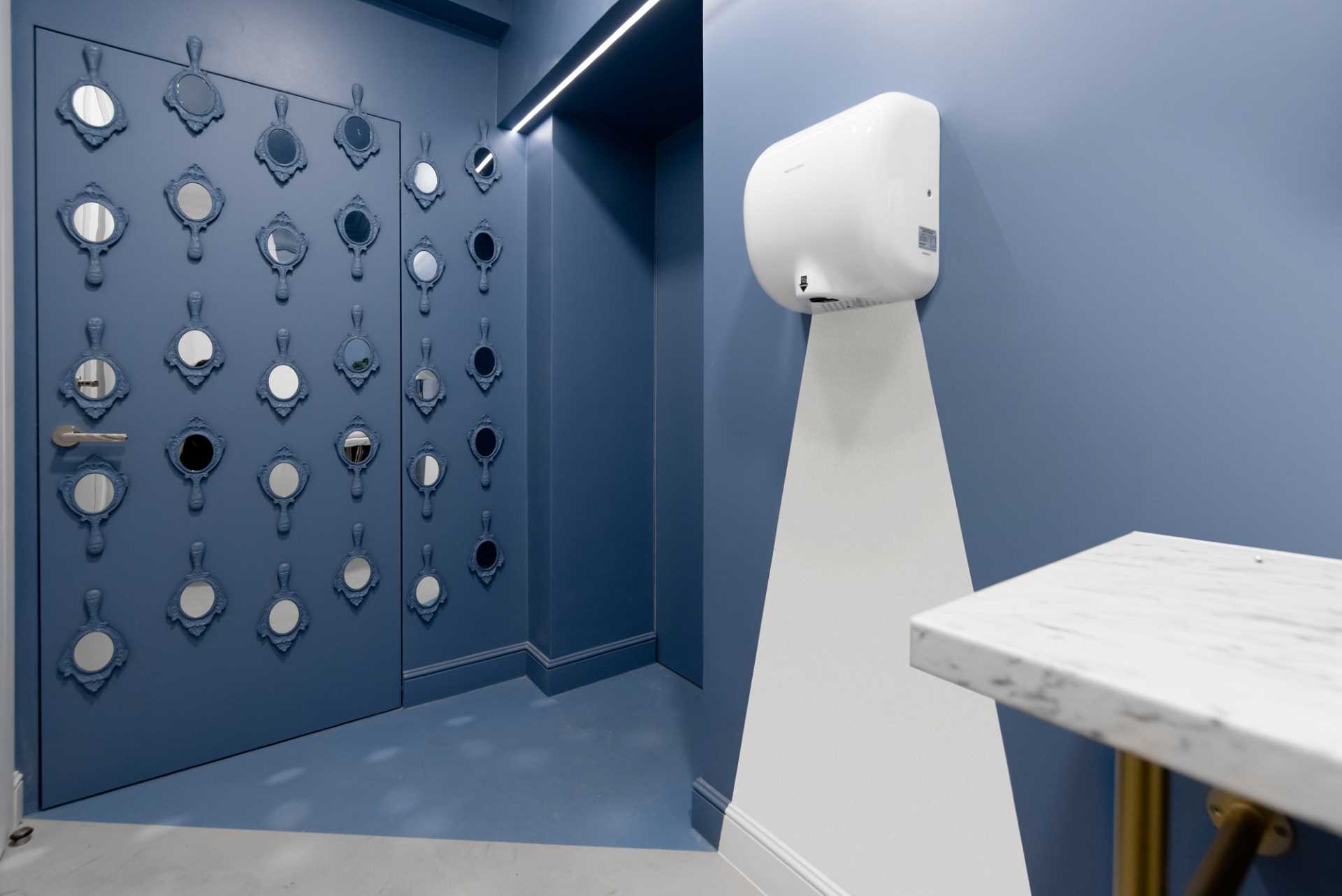 In this office bathroom, the blue walls are accented by mirrors, and for a quirky touch, the hand dryer looks like it has light coming out from it, but it's just a painted white accent that lines up with the floor design.