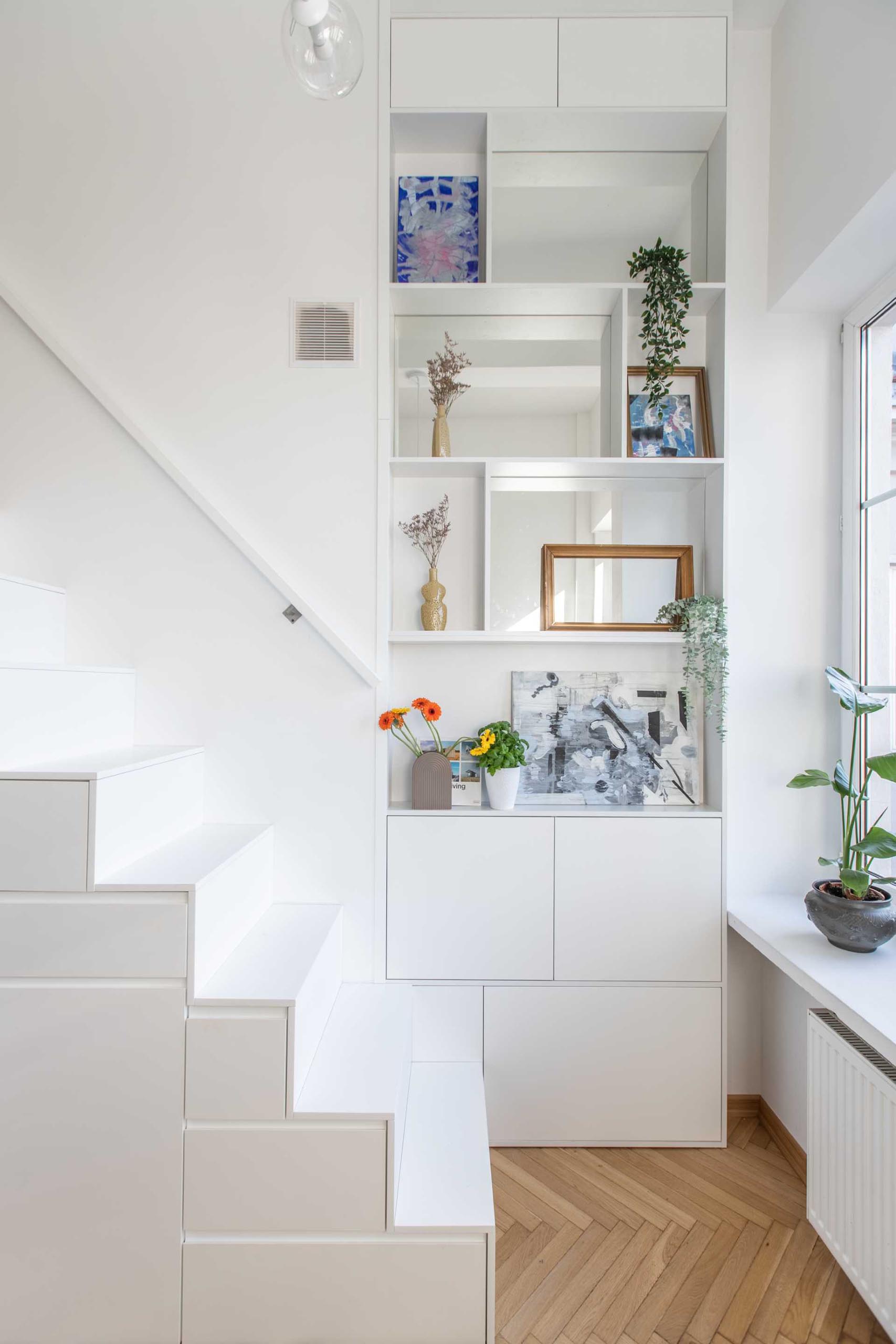 At the bottom of these stairs in a small apartment, there's a floor-to-ceiling shelving unit that includes storage and mirrors.