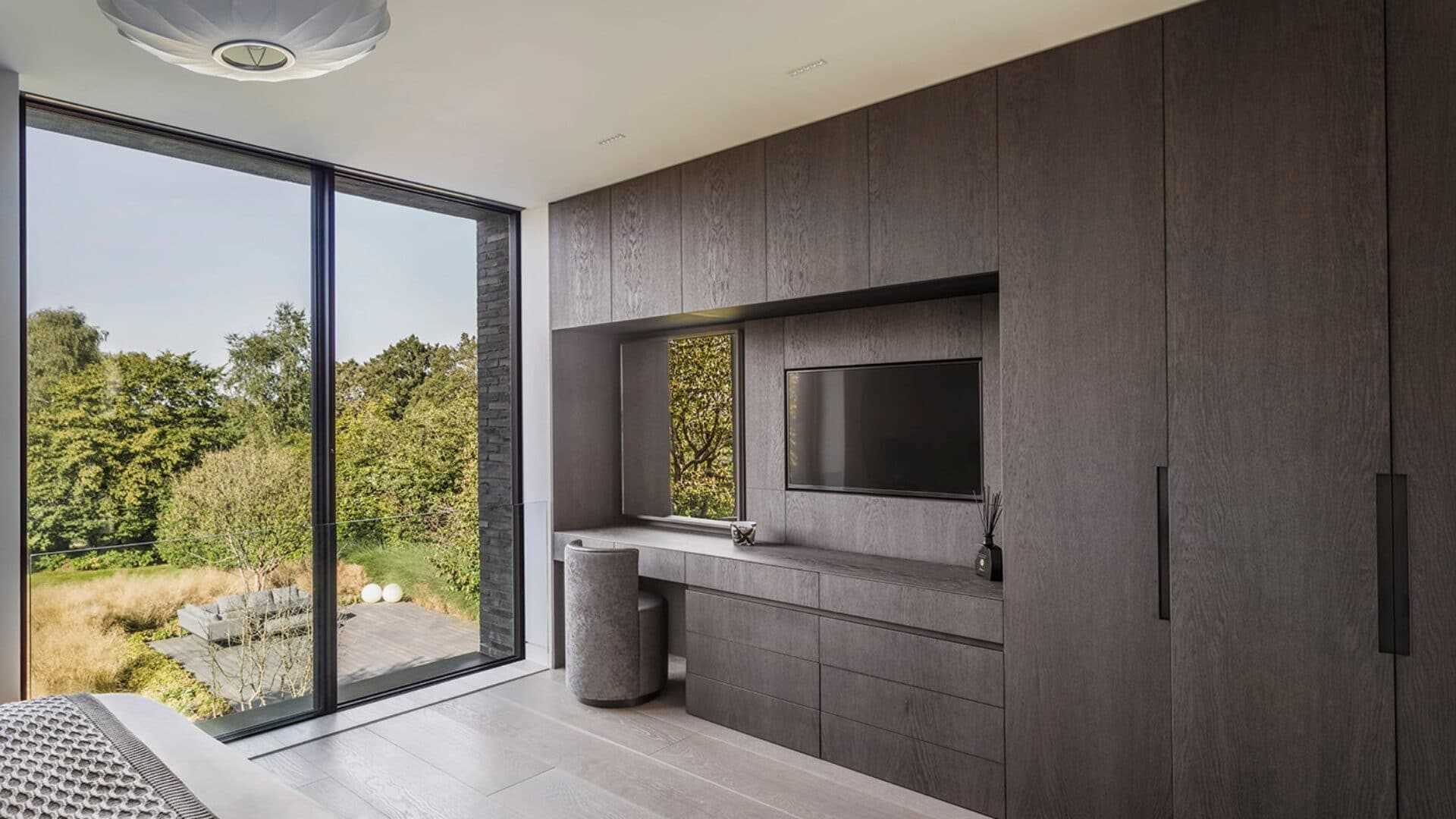 In this modern bedroom, large floor-to-ceiling windows overlook the garden, while built-in closets include a vanity area and built-in TV.