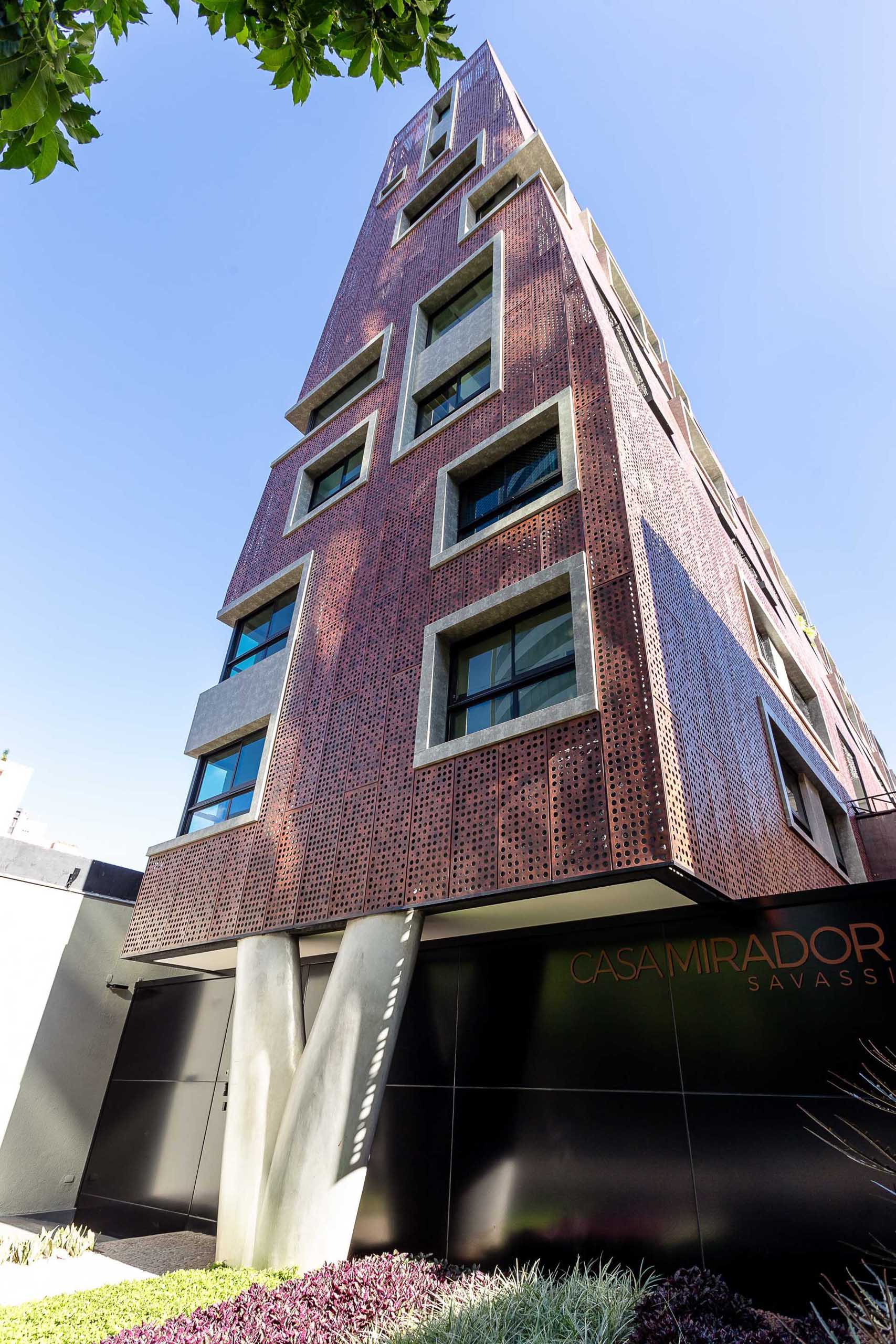 The facade that covers this modern residential building is made from perforated aluminum panels, each painted in an earthy reddish sepia tone.