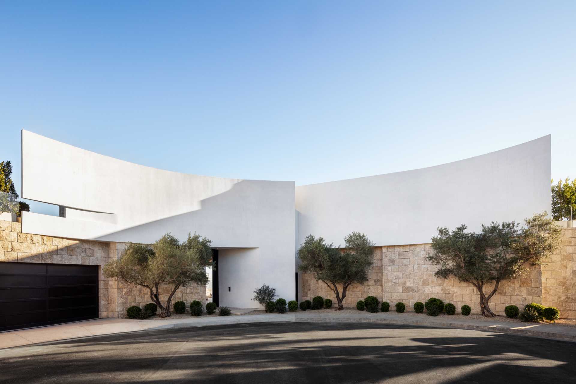 Upon arriving at the home, there's large curved white walls accented by stone and olive trees, with the entryway highlighted by lighting.