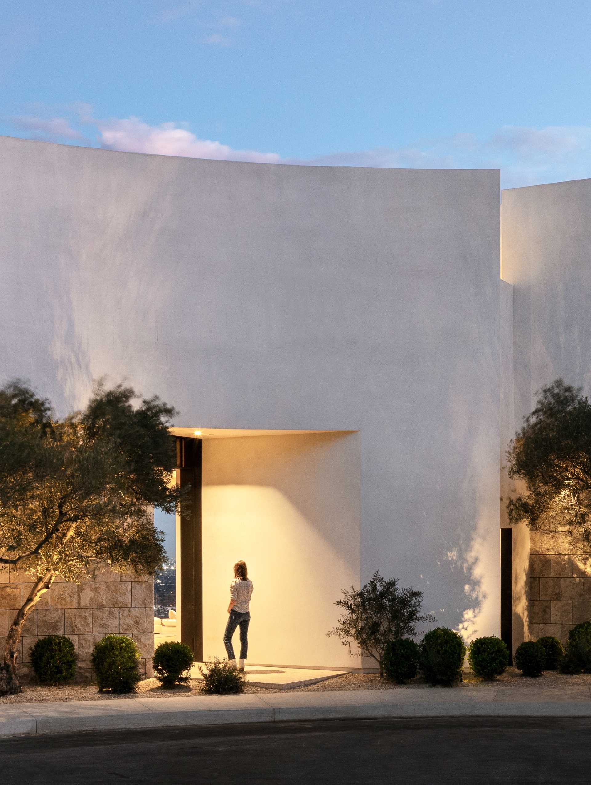 Upon arriving at the home, there's large curved white walls accented by stone and olive trees, with the entryway highlighted by lighting.
