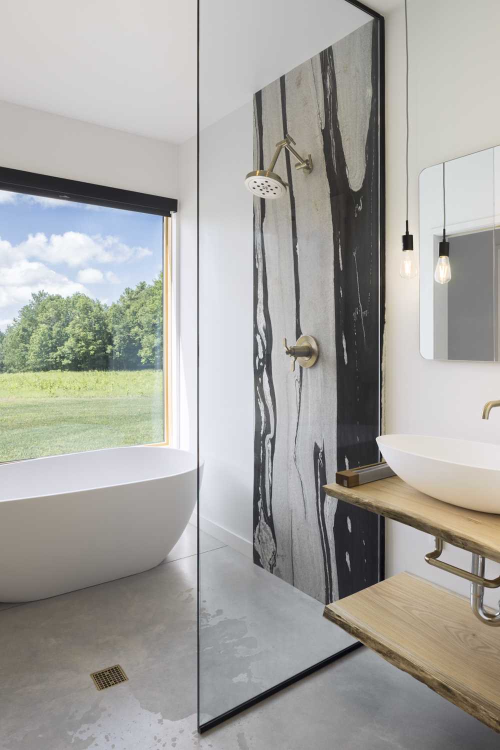 In this modern bathroom, locally sourced oak slabs form the vanity, while brass fixtures from Brizo are water-conservation conscious, and a large window provides unobstructed views of the field.