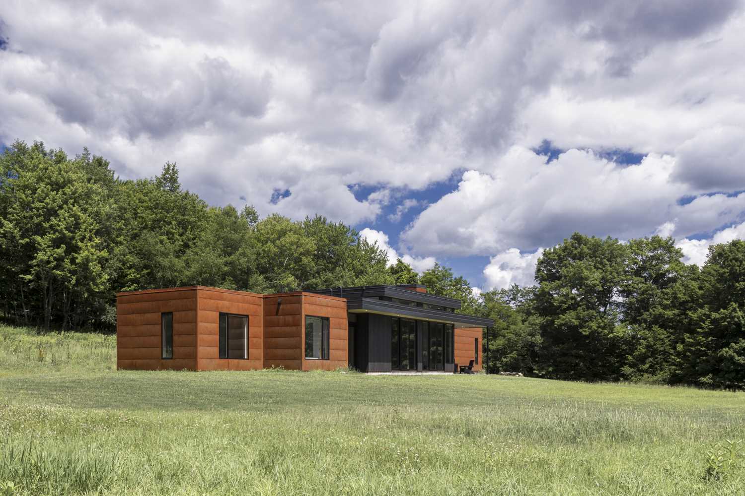A modern house in a field, has a weathered steel exterior and black-stained pine siding.