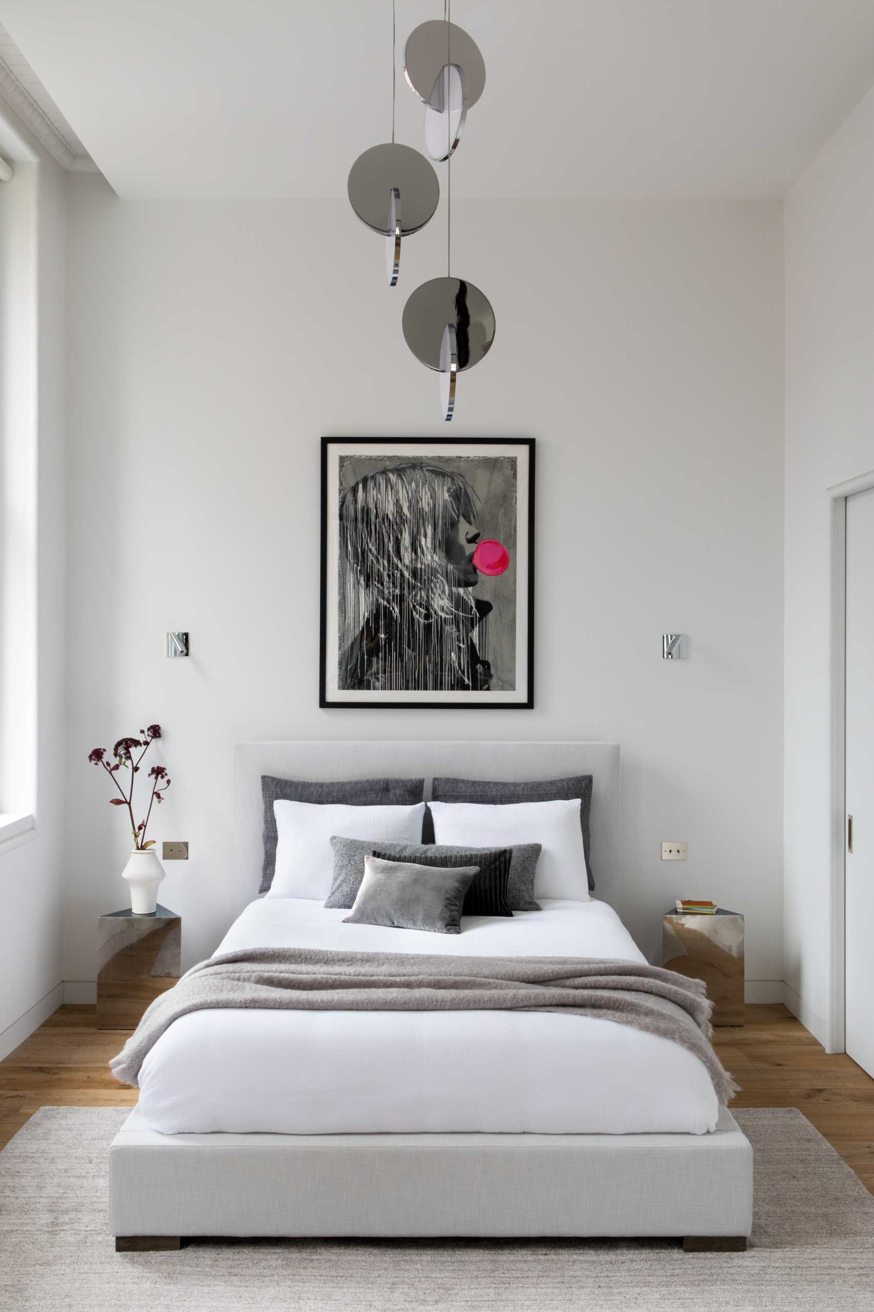 This modern bedroom, located by the windows, includes an Eclipse Chandelier by Lee Broom, a Pacha Lounge Chair by Pierre Paulin, and a Bubble Gum Girl Painting by Hijack.