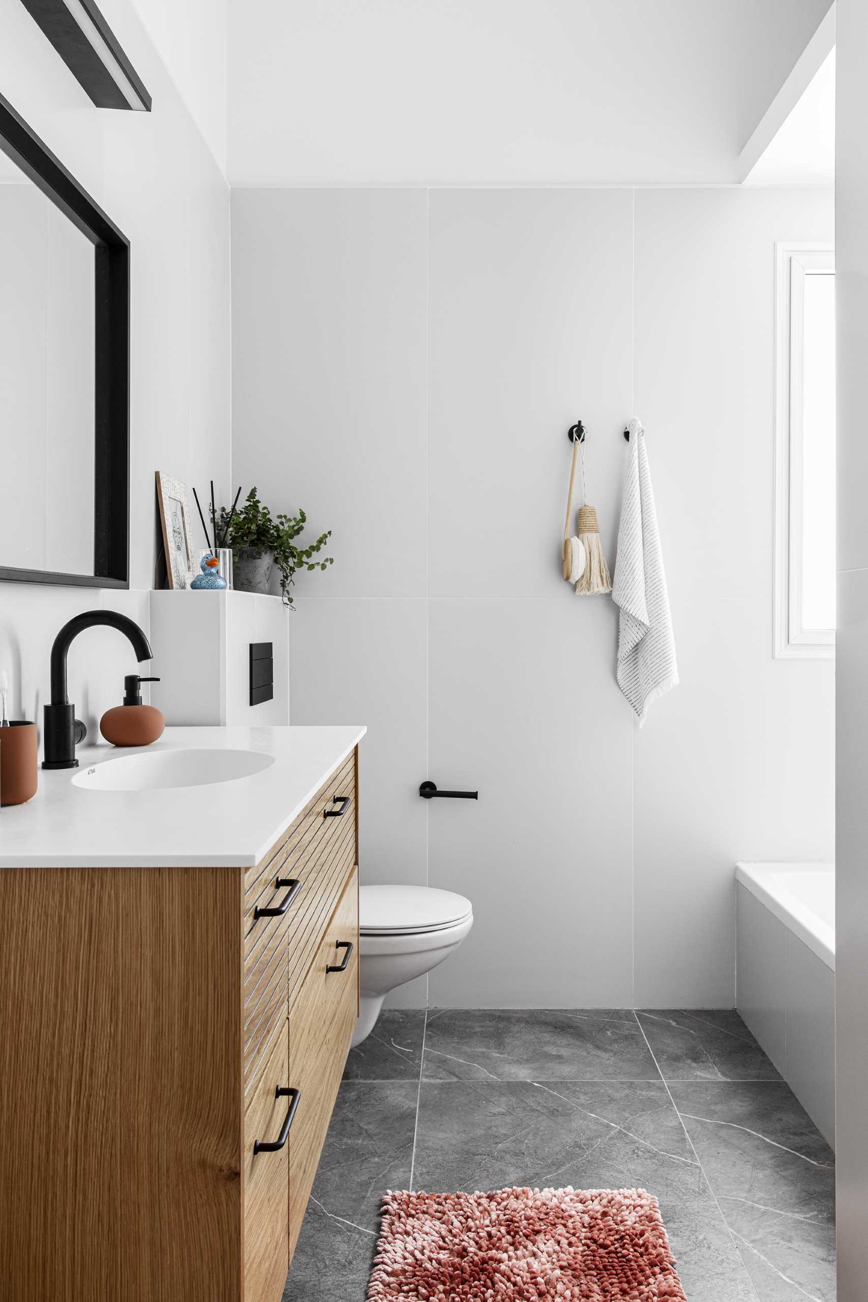 Neste banheiro moderno, há uma pia de madeira flutuante, um espelho quadrado de moldura preta, um par de ganchos de parede, azulejos de grande formato e um combo banheira/chuveiro.  Escondidas da vista estão as máquinas de lavar.