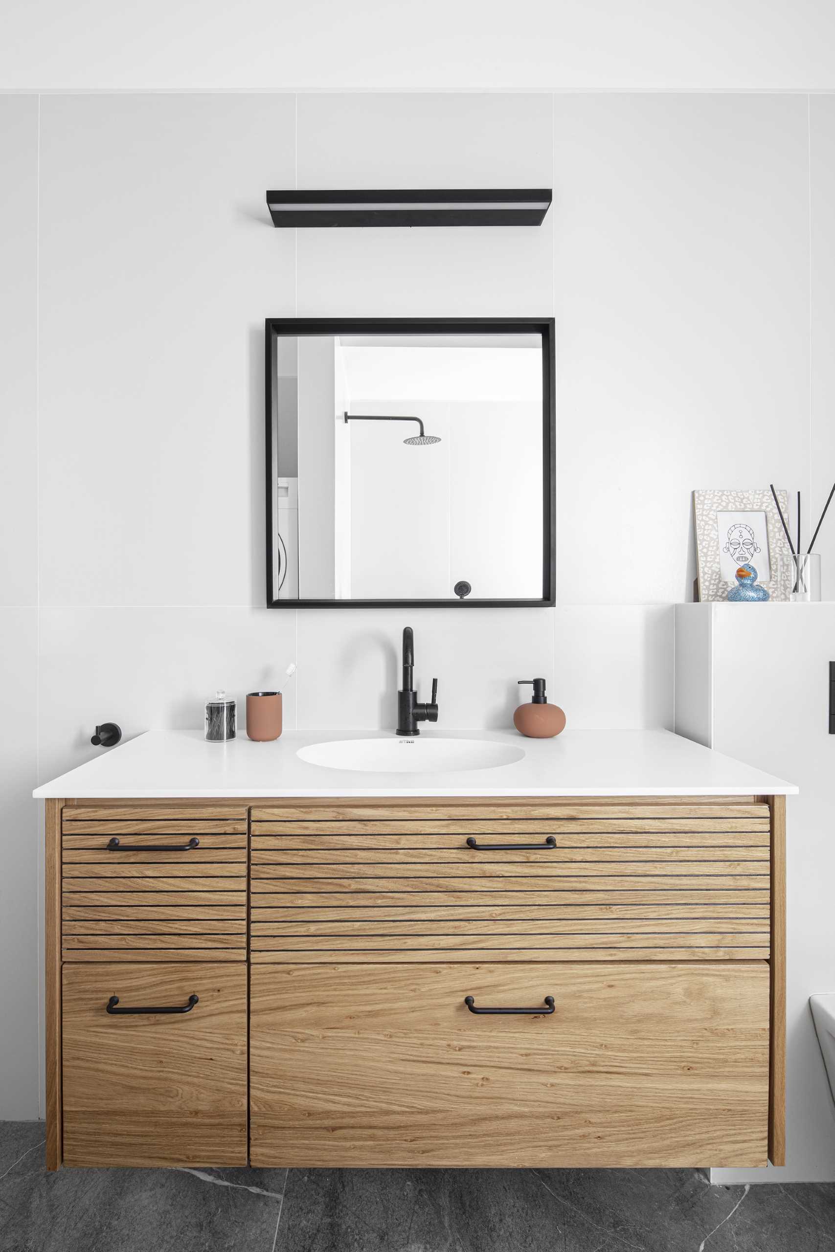 Neste banheiro moderno, há uma pia de madeira flutuante, um espelho quadrado de moldura preta, um par de ganchos de parede, azulejos de grande formato e um combo banheira/chuveiro.  Escondidas da vista estão as máquinas de lavar.